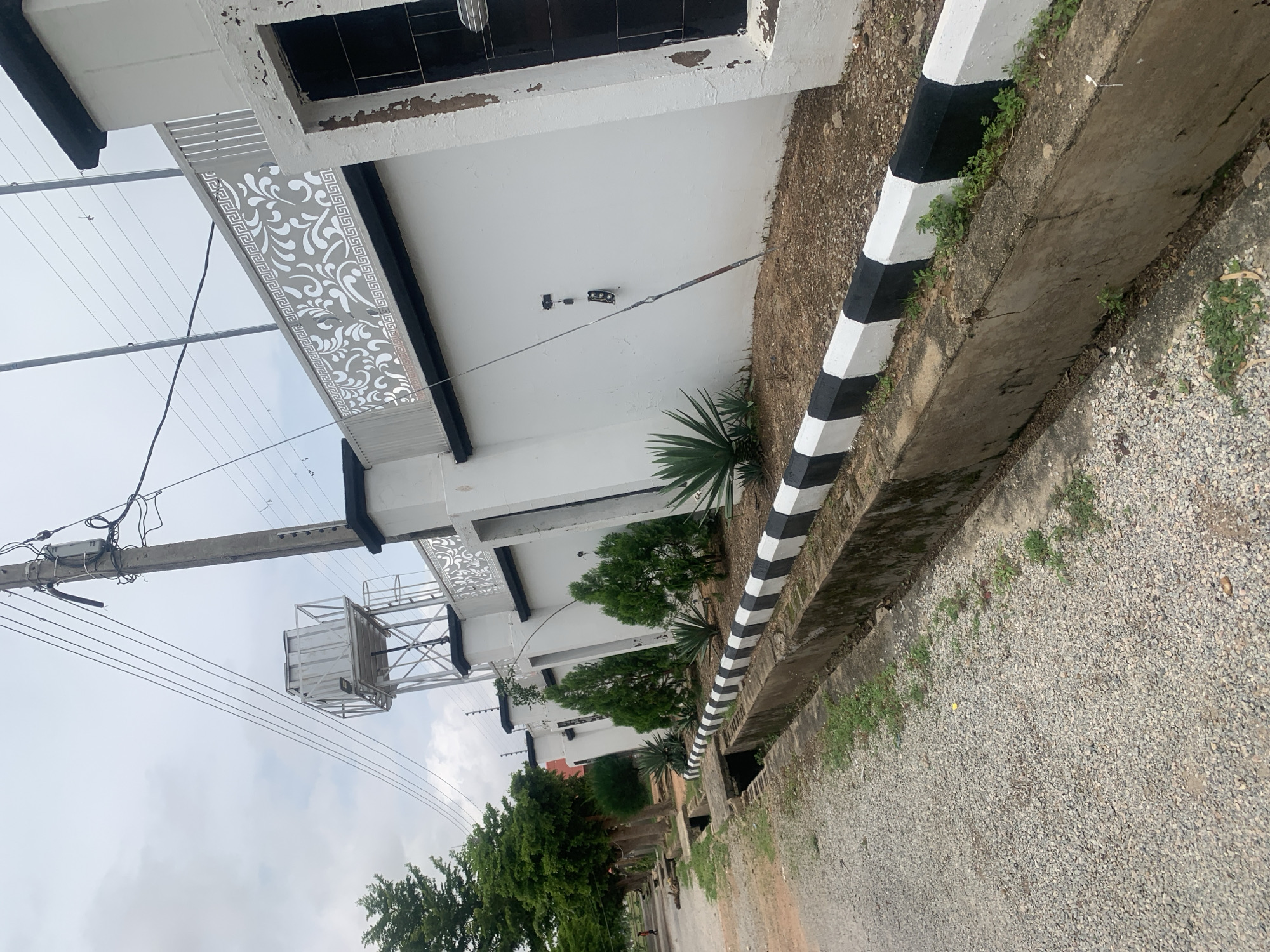 The image depicts a street scene in Jauro Yinu, Nigeria. In the foreground, a gravel road runs alongside a concrete drainage ditch with a black and white painted curb. The ditch contains some sparse vegetation and debris. Beyond the ditch, a white wall with decorative black and white elements runs along the perimeter of a property. Several evergreen trees and agave plants are situated in a landscaped area behind the wall. A utility pole with electrical wires and a water tank on a metal structure are visible in the background, extending into the cloudy sky. The overall scene suggests an outdoor residential or commercial area in a Nigerian town.