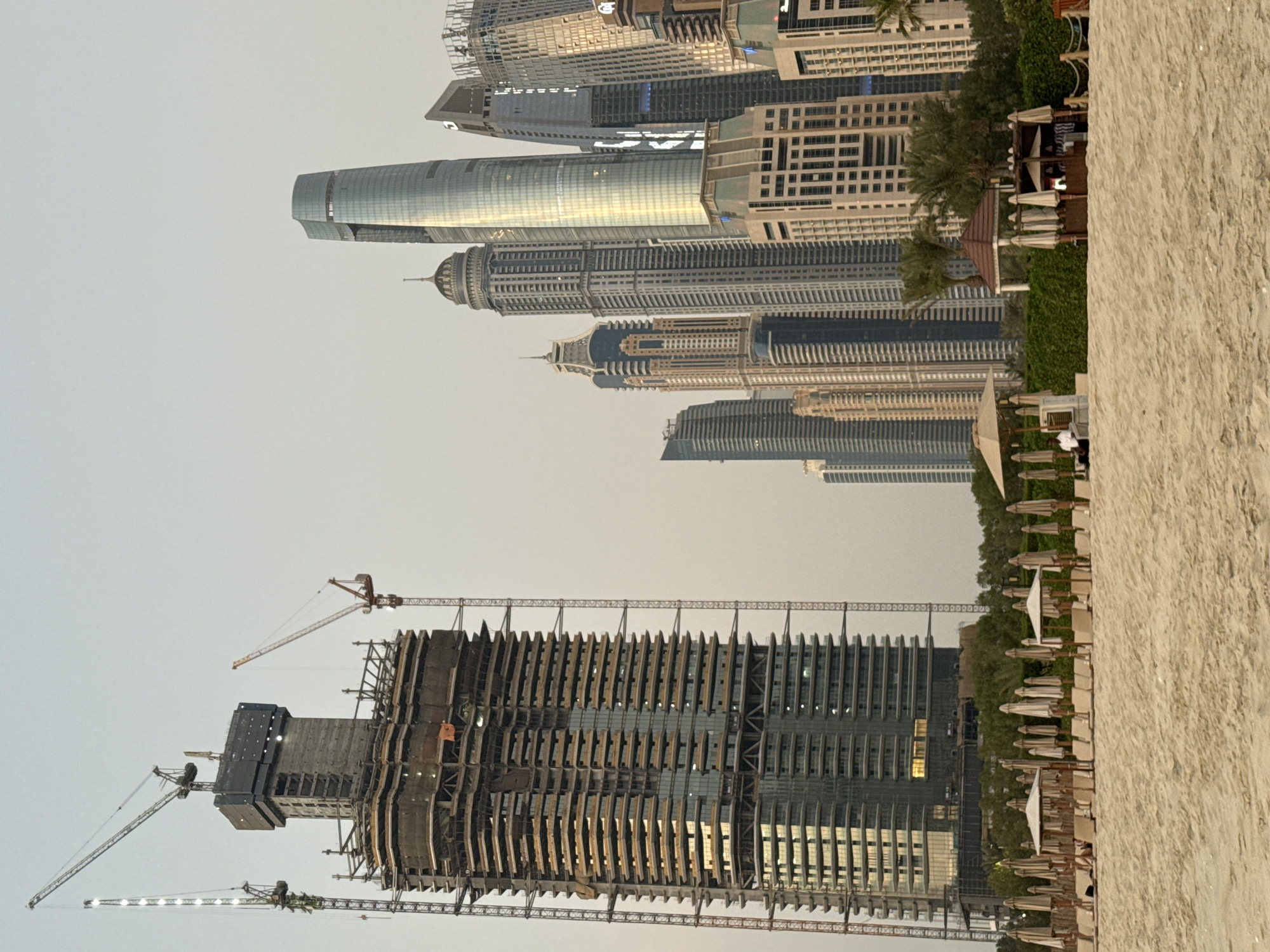 The image depicts a construction site and a modern cityscape in Dubai, United Arab Emirates, with a beach in the foreground. A very tall skyscraper, under construction, dominates the left side of the frame, with multiple cranes surrounding it. To the right, a cluster of completed skyscrapers, characterized by their modern architectural designs and glass facades, are visible, reflecting the soft daylight. The beach in the foreground is sandy, with rows of closed umbrellas and lounge chairs, suggesting an unoccupied or off-season resort area. Behind the beach, a line of palm trees and dense green hedges separates it from the skyscrapers. A small, open-air structure with a red roof and white canopies is situated among the trees. One person, dressed in white, is visible near this structure. The sky is a pale, overcast gray, indicating diffused light conditions.