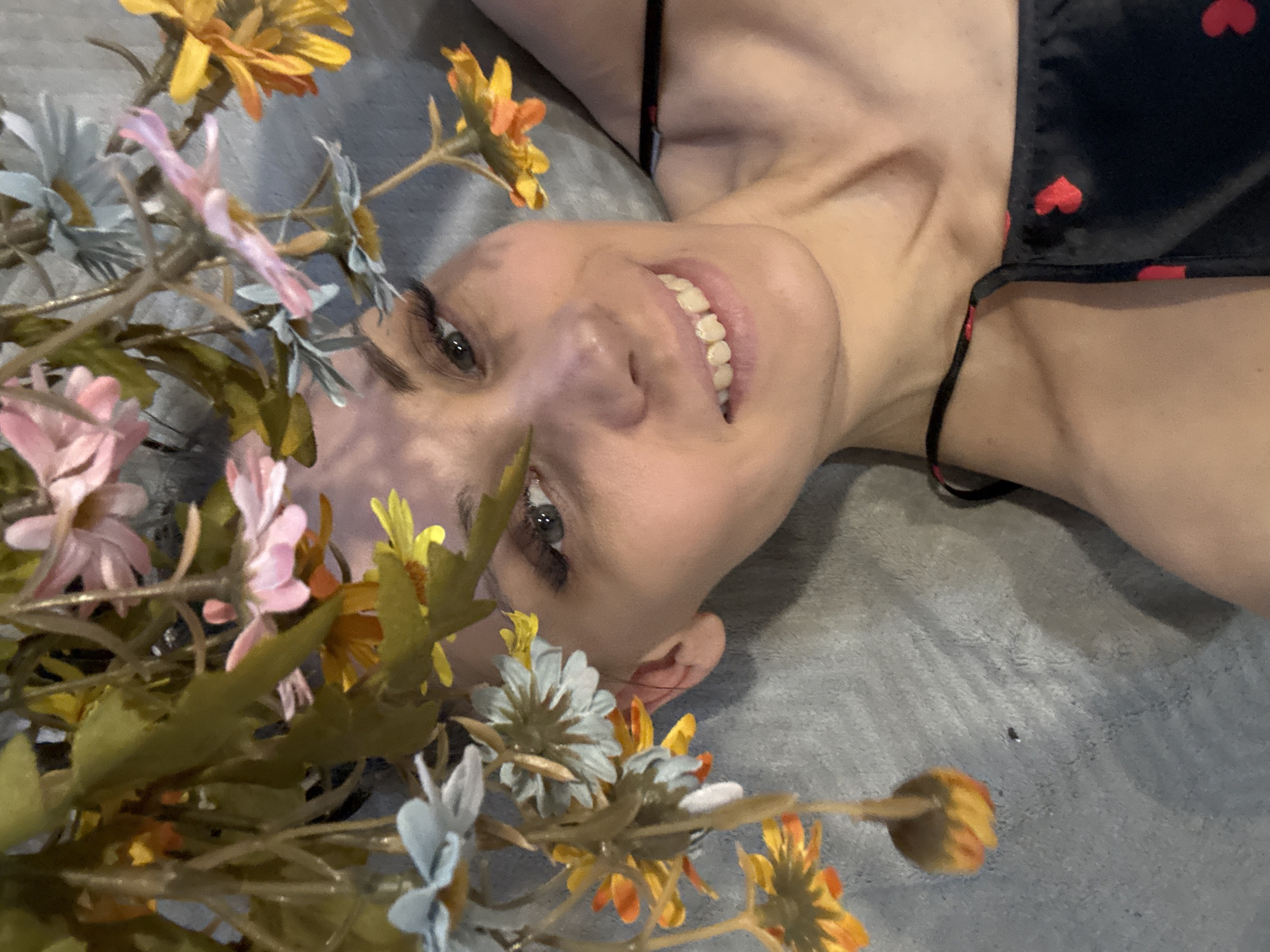 A woman is lying on a gray blanket, smiling. She is wearing a black top with red hearts. Artificial flowers in shades of pink, yellow, orange, and blue are positioned in front of her face. The location is unknown and cannot be determined from the image.