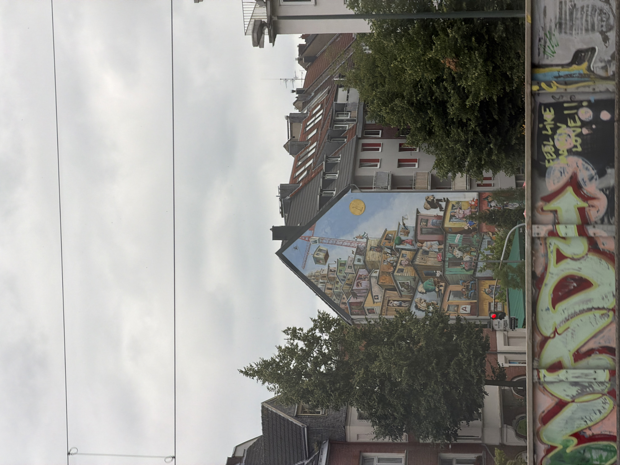 The image depicts a section of a building in Dusseldorf, Germany, adorned with a vibrant mural. The mural showcases a fantastical, stacked city composed of numerous small buildings, with cranes and various figures integrated into the scene, creating a sense of whimsical chaos. In the foreground, a graffiti-covered wall displays colorful tags and lettering. The sky is overcast, and trees are visible in front of the building. A traffic light with a red signal is also present. The overall scene captures an urban environment with a focus on street art and architecture.