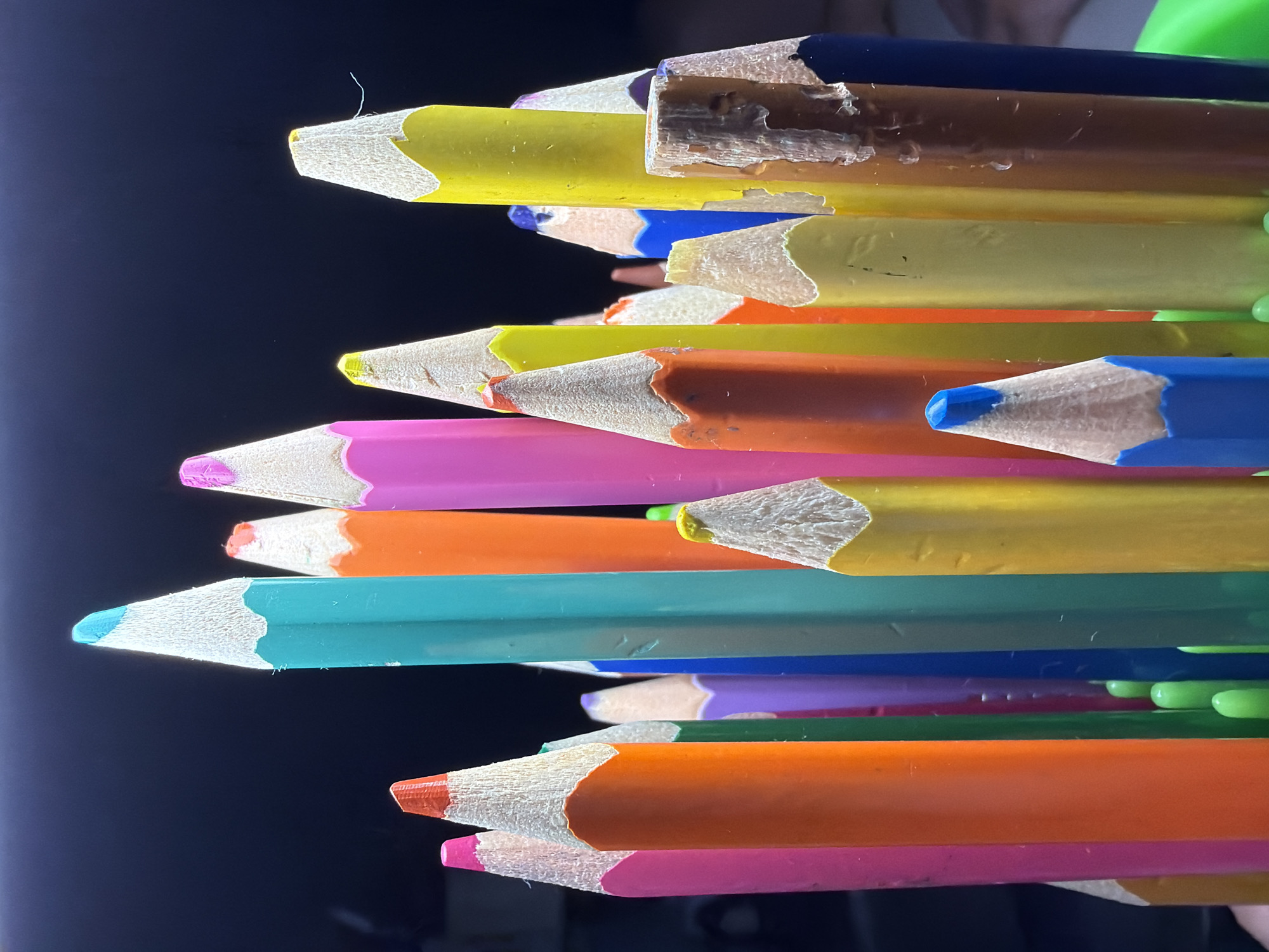 A close-up, vertically-oriented photograph shows a stack of colored pencils against a blurred dark background. The pencils are arranged haphazardly, with their sharpened points facing the viewer's left. The pencils are various colors, including yellow, brown, blue, orange, pink, green, purple, and teal. The points of the pencils show the exposed wood and colored lead. A hair or thin fiber is visible near the top of the image, extending from a yellow pencil. The background is dark and out of focus. The setting cannot be determined, and there is no visible evidence of the location being Votkinsk, Russia. No people are visible.