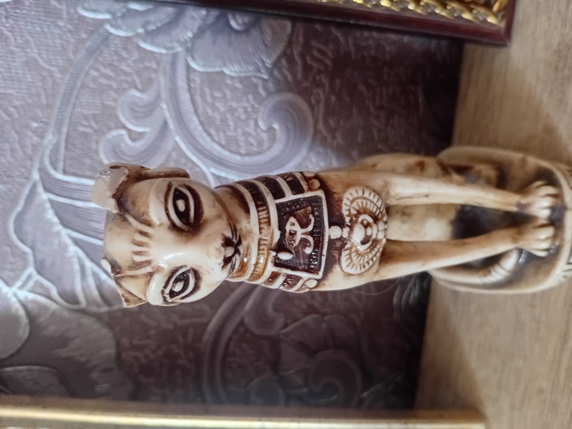 This is a close-up, eye-level, indoor photograph of an Egyptian-style cat figurine. The figurine is made of a light-colored material, possibly resin or carved bone, and has dark brown painted details. It depicts a seated cat with large, almond-shaped eyes outlined in dark brown, a small nose, and painted lines on its forehead and cheeks. The cat wears an ornate collar with horizontal bands and a central square emblem containing hieroglyphic-like symbols and a circular motif below it. The figurine is positioned on a wooden surface, likely a shelf. The background consists of wallpaper with a raised, ornate floral pattern in muted tones of gray and beige. To the left of the figurine, a thin, gold-colored cylindrical object is partially visible, possibly part of a frame or stand. To the right, a portion of a dark red, ornate frame is visible. The lighting is soft and diffused, casting subtle shadows. The scene is static and appears to be a decorative display. The location is identified as Курманаевка, Russia.