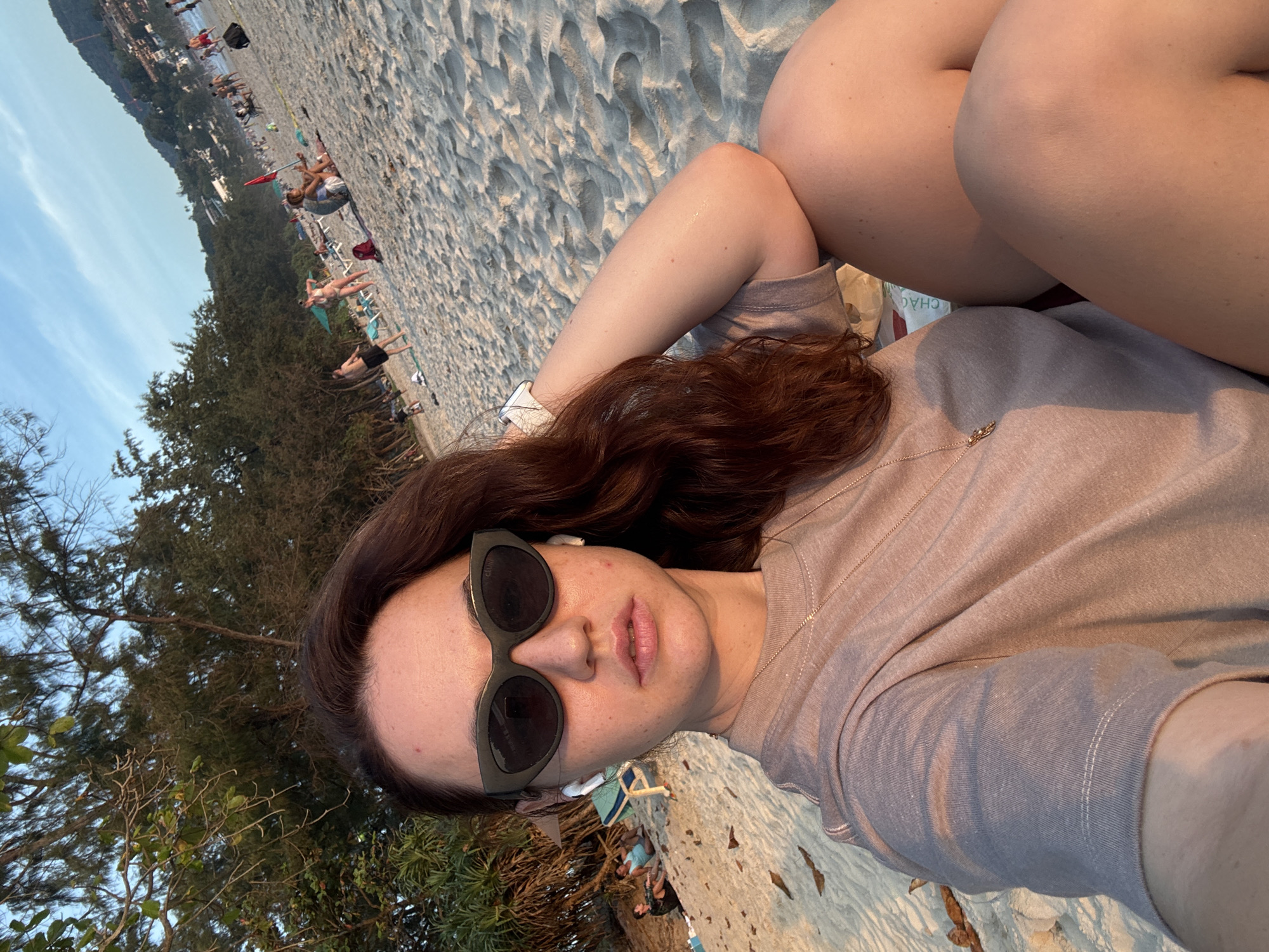 A woman is sitting on a sandy beach, with trees and the ocean in the background. The woman is wearing sunglasses, a t-shirt, and a necklace. She has earbuds in her ears. The beach is populated with other people, some of whom are swimming or sunbathing. The sky is clear and blue, with a few clouds. The overall mood of the image is relaxed and casual.