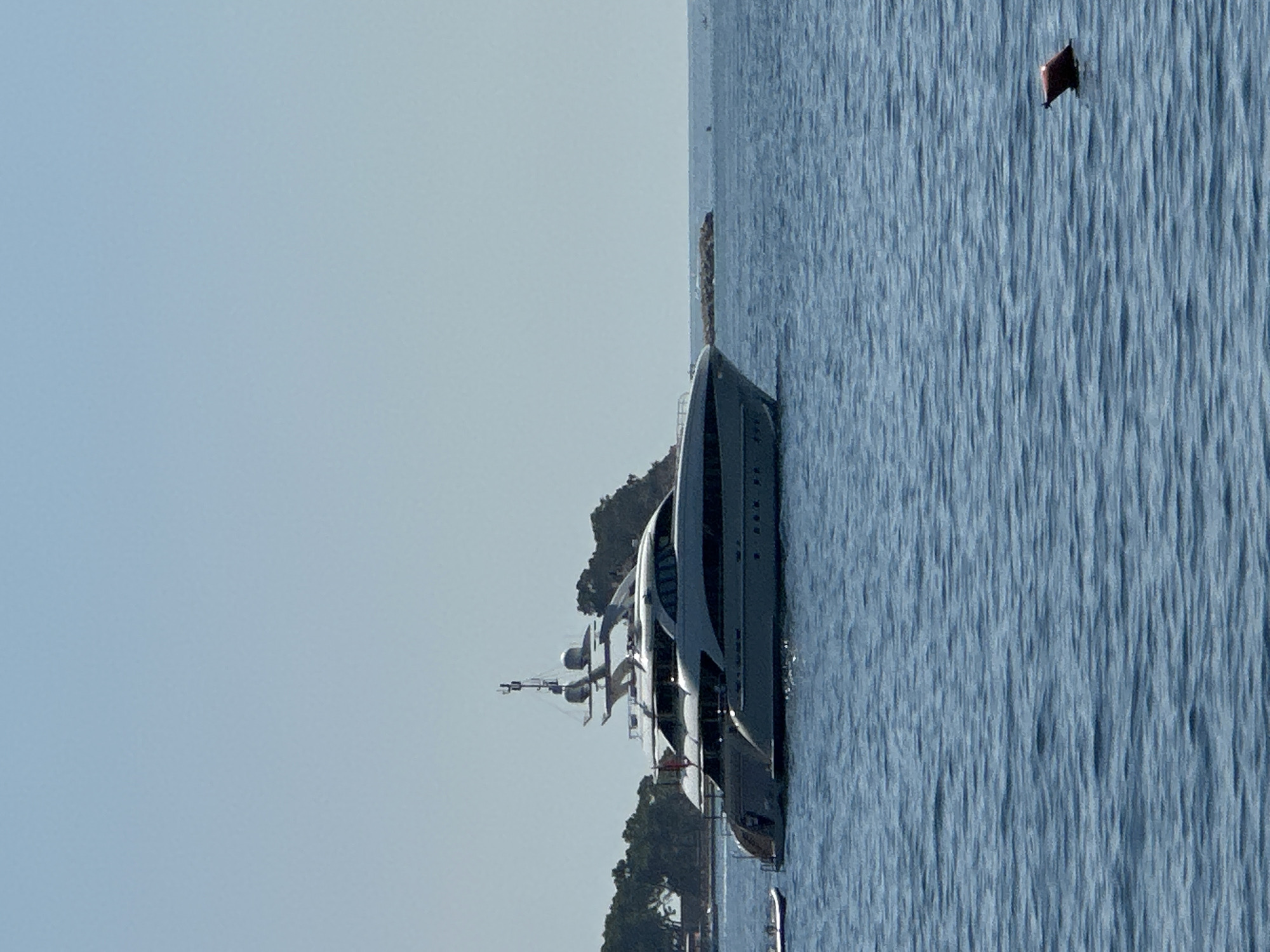A large white motor yacht is anchored in the blue water of the Adriatic Sea, near the coast of Budva, Montenegro. The yacht has multiple decks and appears to be luxurious, with a sleek, modern design. In the background, a tree-covered island and a rocky outcrop are visible. The sky is clear and blue. A small, inflatable boat is tied to the stern of the yacht. A red buoy is floating in the water to the right of the frame. The sea is calm with gentle ripples.