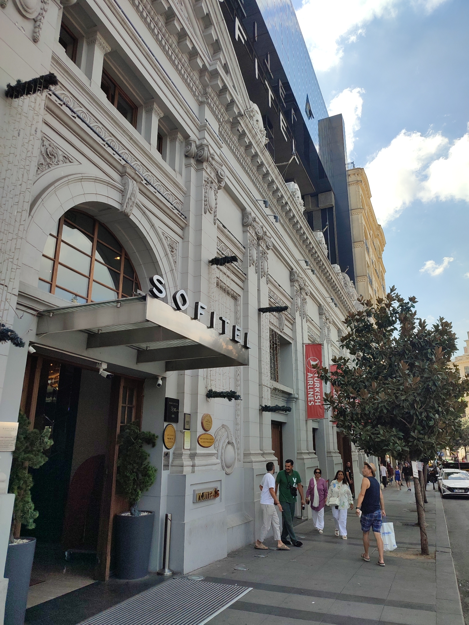 The image shows the exterior of the Sofitel Istanbul Taksim hotel in the Beyoğlu district of Istanbul, Turkey. The hotel's facade is predominantly white with ornate architectural details, including columns and decorative moldings. The "SOFITEL" logo is prominently displayed above the entrance. To the right of the hotel, a modern, darker building with a glass facade rises high into the sky. A red banner with the Turkish Airlines logo hangs on the hotel's exterior. Several people are walking on the sidewalk in front of the hotel. A large, leafy tree is positioned to the right of the hotel entrance. The sky is blue with scattered white clouds. There are potted plants and outdoor seating near the entrance. The street is paved, and a white car is visible further down the street.