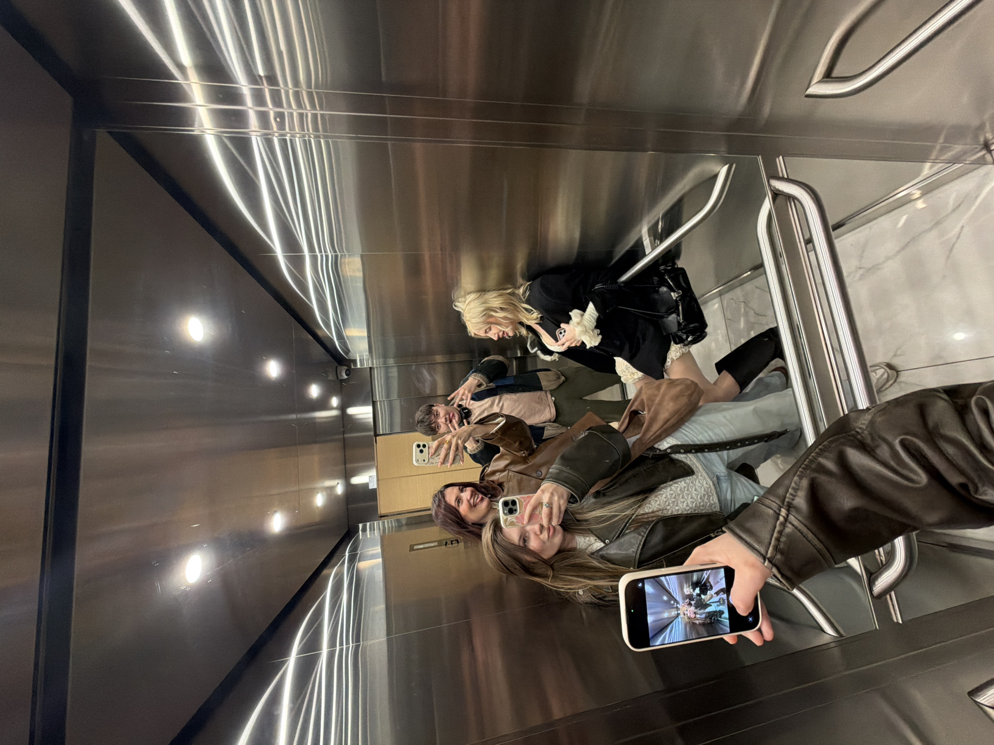 Four people are in an elevator, taking a photo of themselves in the mirrored walls of the elevator. Three people are holding up their phones in order to take a photo of each other. The elevator walls and ceiling are made of brushed metal and feature long fluorescent light fixtures. The floor appears to be white marble. There is a handrail installed at the right corner of the elevator. It is not possible to independently verify that this image was taken in the city of Belgrade, Serbia.