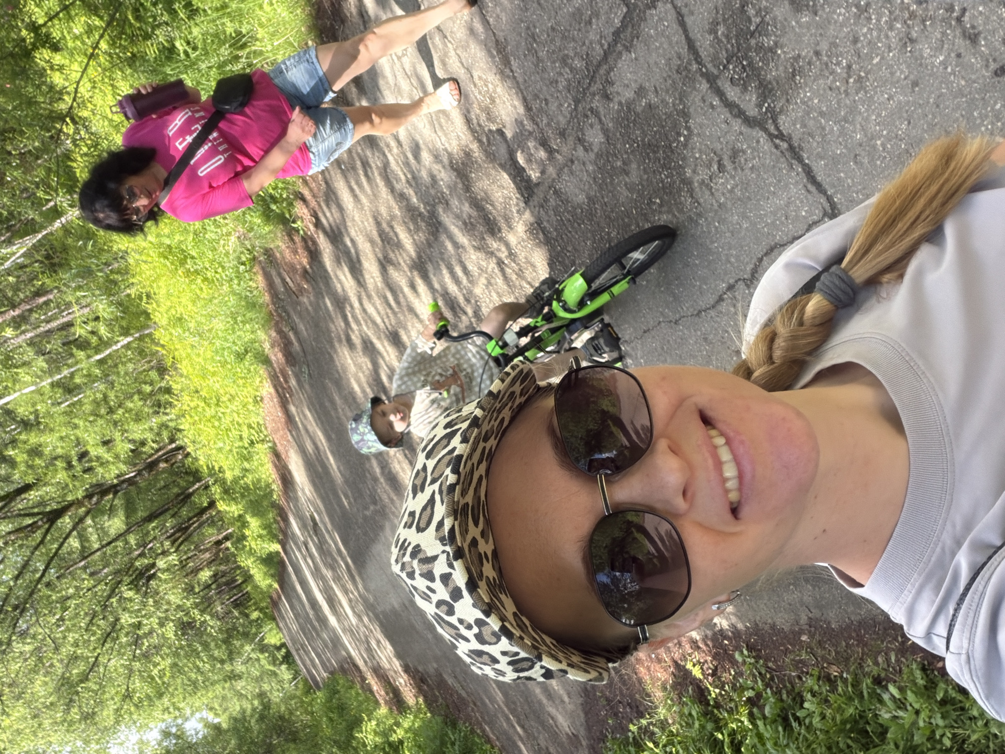 In Кульдур, Russia, on a tree-lined asphalt path, a woman in a leopard-print cap and sunglasses takes a selfie. Behind her, a young child rides a bright green bicycle, while another woman in a pink shirt and denim shorts walks with a purple water bottle in her hand. The scene is bathed in sunlight filtering through the dense green foliage.