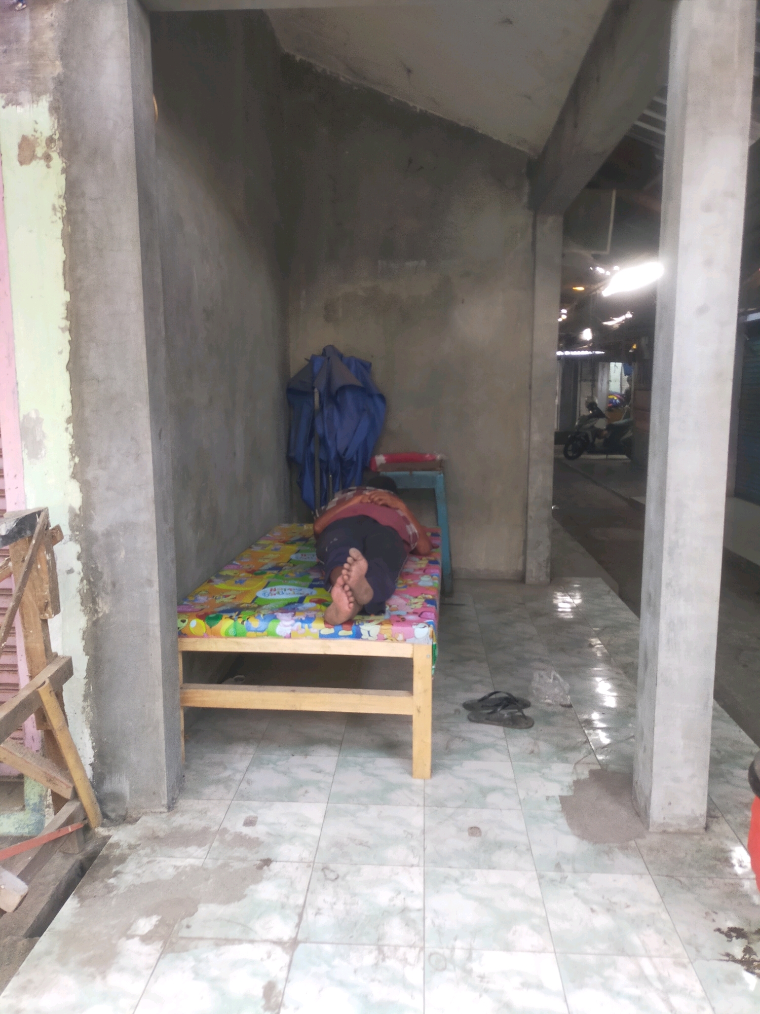 In Bululawang, Indonesia, a person sleeps on a colorful, cartoon-themed mattress atop a simple wooden bed frame. The bed is positioned in an outdoor, covered area with concrete walls and a tiled floor that shows signs of wear and moisture. To the left of the bed, a blue tarp is draped over what appears to be a folding chair. In the background, a narrow walkway leads to an area where a motorcycle is parked. The overall scene suggests a humble, functional living or resting space, likely part of a shop or market stall.