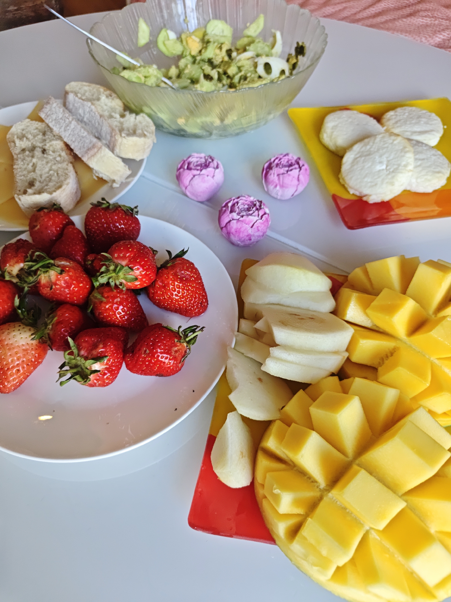 The image presents a tabletop arrangement of food items on a white table surface. A clear glass bowl contains a salad of cucumbers and what appear to be avocado and hard-boiled eggs, with a serving spoon present. Adjacent to this, a white plate holds slices of bread and cheese. A separate white plate is piled with fresh strawberries. A brightly colored cutting board displays slices of peeled pears and diced mango. A yellow and red plate contains rounds of white cheese. Three small, purple ornamental balls are positioned between the salad bowl and the other dishes. A pink knitted blanket is visible in the background. No people are visible in the frame. There is no evidence to support the assertion of this photo being in Sochi, Russia.