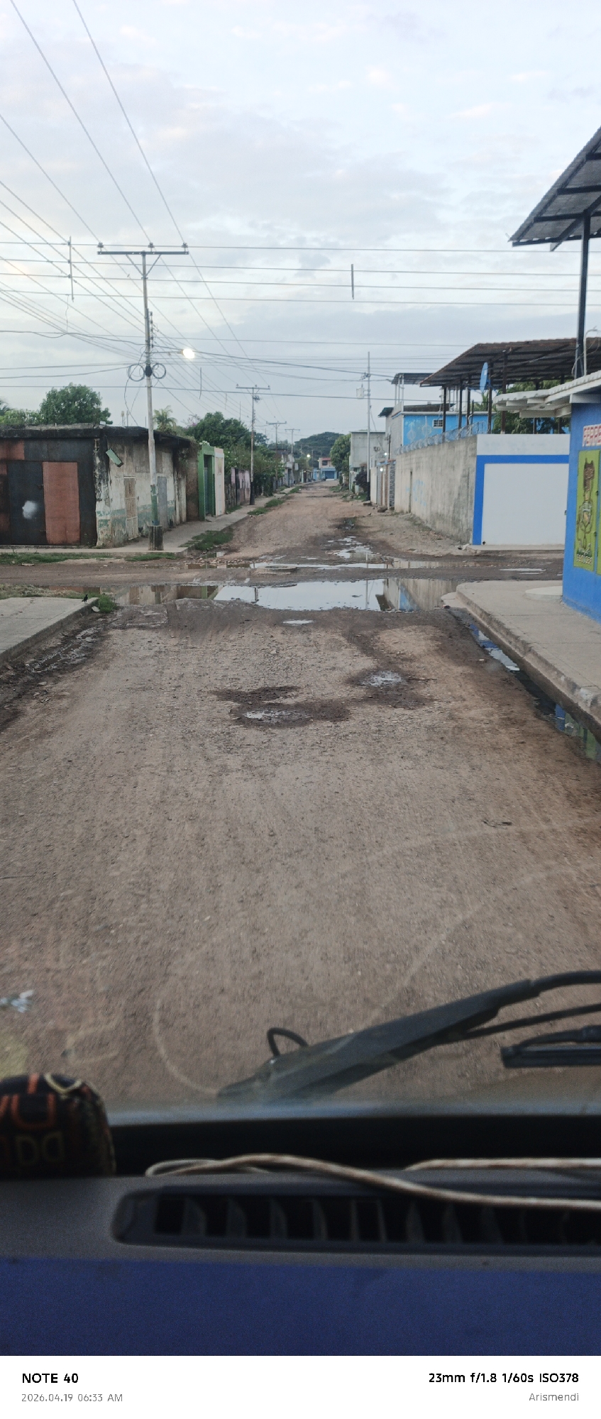 The image depicts a dirt road with numerous potholes filled with water in a low-income neighborhood in Arismendi, Venezuela. The perspective is from inside a vehicle, with part of the dashboard and windshield visible in the foreground. On either side of the road are dilapidated houses and buildings, many with mismatched or unfinished exteriors. Utility poles with tangled wires line the street, contributing to the unkempt appearance. The sky is overcast, indicating either early morning or late afternoon lighting conditions. The timestamp indicates the photo was taken in 2026.