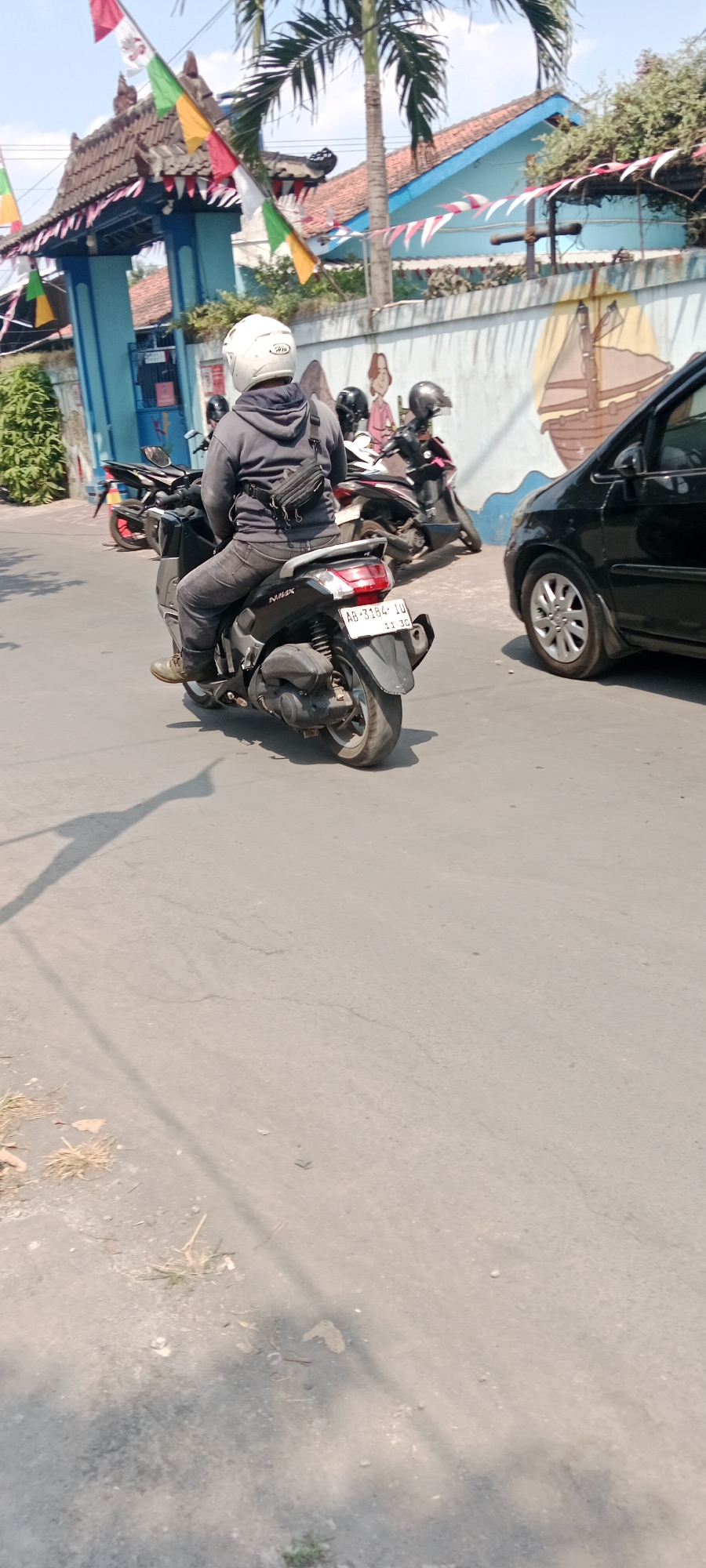 The image shows a street scene in Purwomartani, Indonesia. In the foreground, a person is riding a black motorcycle. The rider is wearing a gray hoodie, dark pants, and a white helmet. They have a black fanny pack around their waist. The motorcycle has a license plate that reads "AB 3184 ID". Behind the motorcycle, another black motorcycle is parked. To the right, the rear section of a black car is visible. In the background, there is a blue building with a traditional roof and flags. A mural of a sailboat is painted on a wall behind the motorcycles. A palm tree is also visible. The ground is paved with asphalt, and there are some dry leaves and grass on the side of the road. The lighting suggests it is daytime with a bright sun, casting shadows on the road.