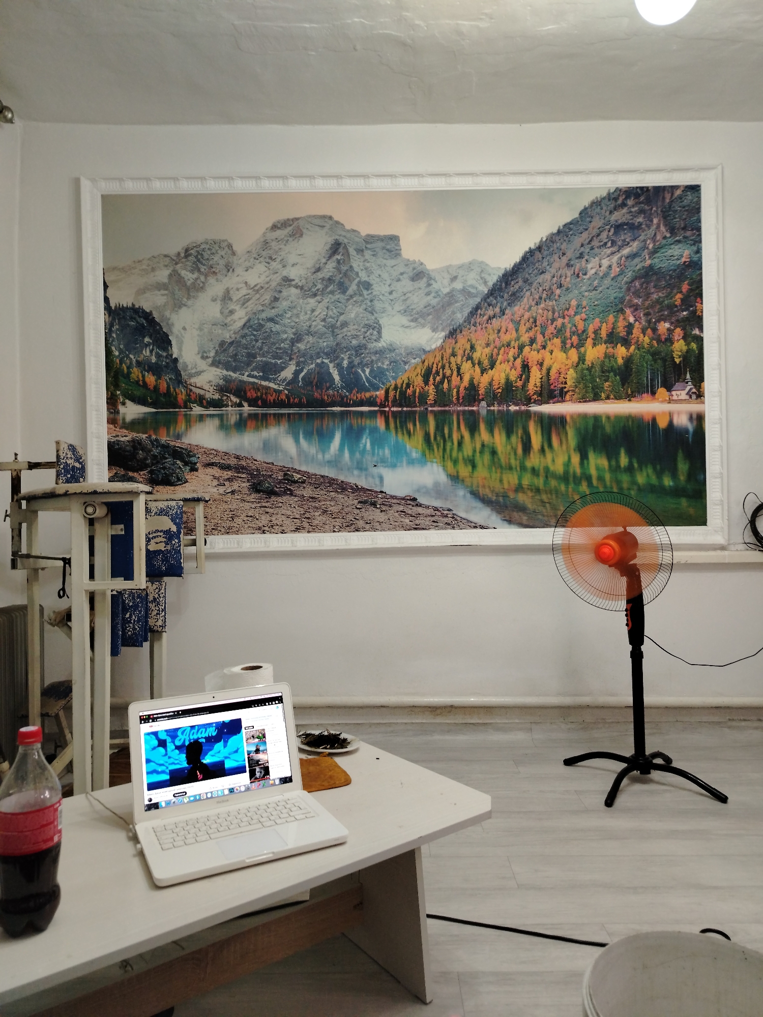 The image depicts an indoor scene. Dominating the background is a large panoramic mural of a mountainous landscape with a lake, featuring autumn foliage. In the foreground, a white table holds an open white laptop displaying a video with the title "Adam" and a silhouette of a person. To the left of the laptop is a bottle of dark soda. A roll of toilet paper is positioned behind the laptop. To the right of the table, a black oscillating fan with an orange light in the center is in operation. A white wooden structure with blue fabric panels stands to the left of the mural. The room has white walls and a light-colored floor. The scene suggests a casual living or work space, possibly a living room or a personal office. There is no indication of the location being specifically in Bishkek City, Kyrgyzstan.
