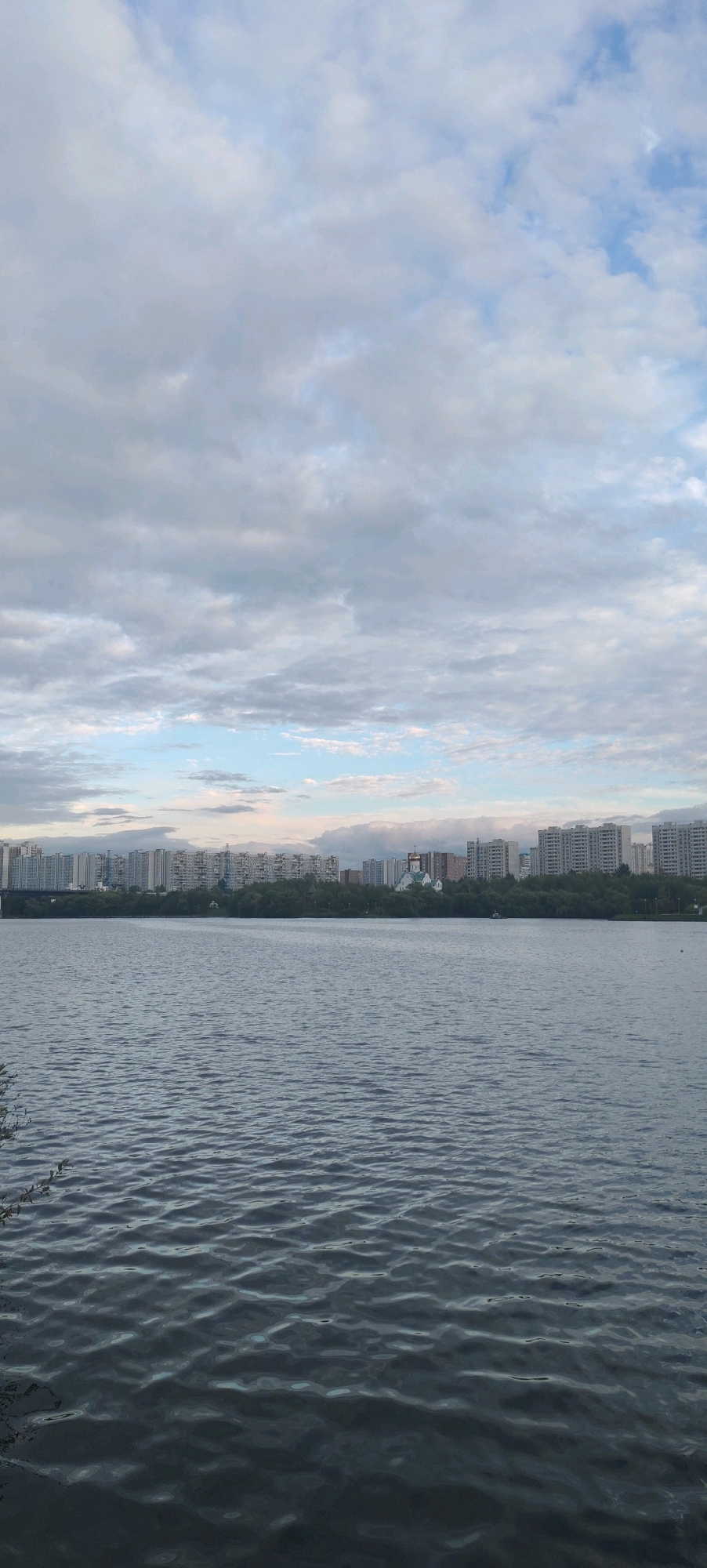 The image shows a panoramic view of a wide river with a city skyline in the background. The river occupies the lower half of the frame, with gentle ripples on its surface. In the middle ground, a line of trees forms a dense green barrier along the riverbank. Behind the trees, a cityscape with numerous apartment buildings stretches across the horizon. Most of these buildings are tall and modern, characterized by repetitive window patterns. To the right of the cluster of apartment buildings, a prominent church with a golden dome and green roof stands out. The sky above is filled with a mix of gray and white clouds, with patches of light blue visible. The overall impression is of a calm, overcast day in an urban environment with a prominent natural feature. The presence of the church with its distinct architecture and the style of the apartment buildings suggest a location in Russia, likely Moscow.