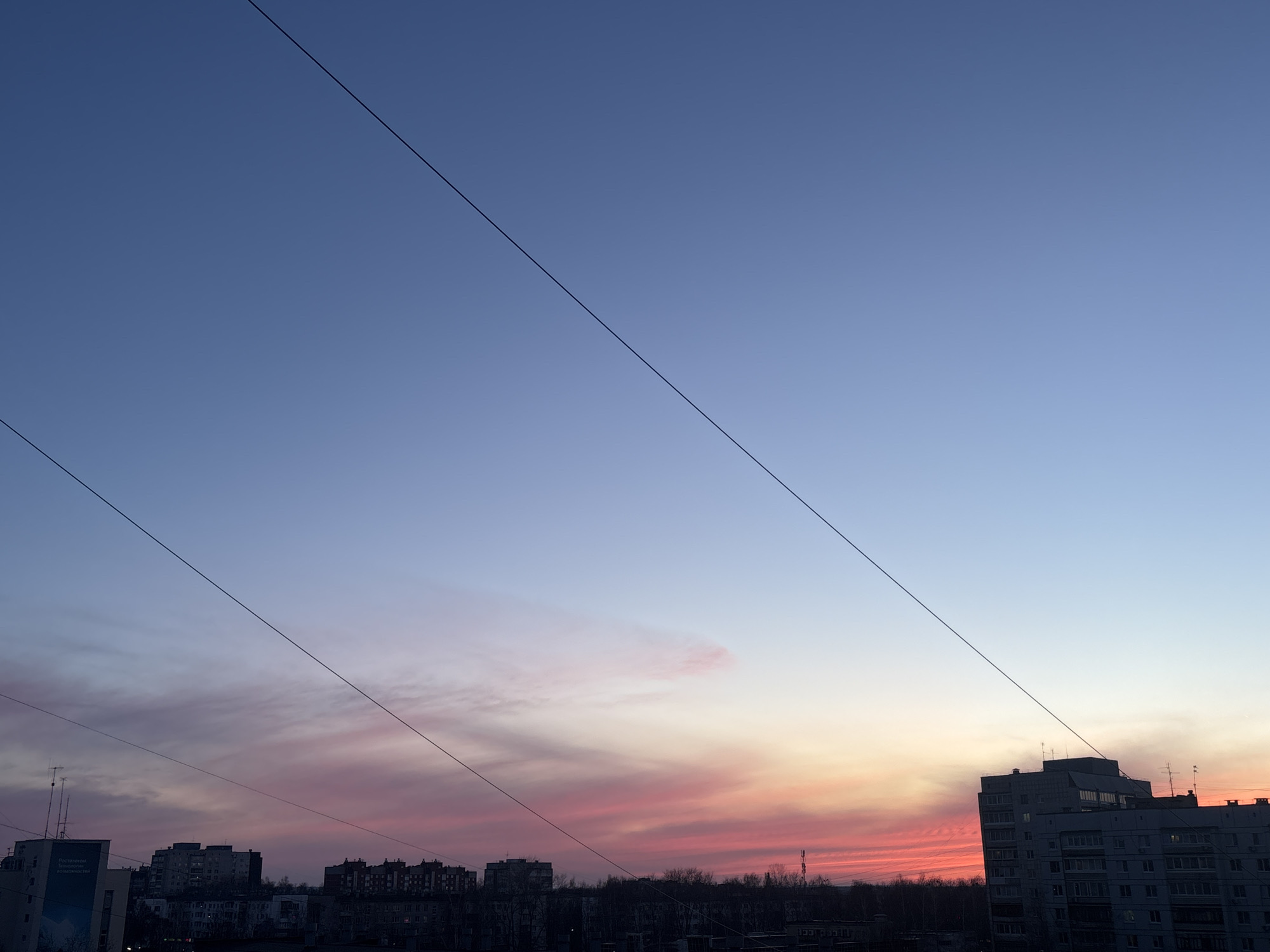 The image captures an urban sunset scene in Chaikovsky, Russia. In the foreground, silhouettes of several multistory buildings dominate the lower portion of the frame, with some buildings displaying visible windows. Dark, leafless trees punctuate the gaps between the buildings. Overhead, three parallel power lines stretch across the frame, intersecting the sky. The sky itself exhibits a gradient of colors, transitioning from a deep blue at the top to hues of pink, orange, and red near the horizon, indicative of a sunset. The overall lighting suggests twilight, with the buildings and foreground elements cast in shadow.