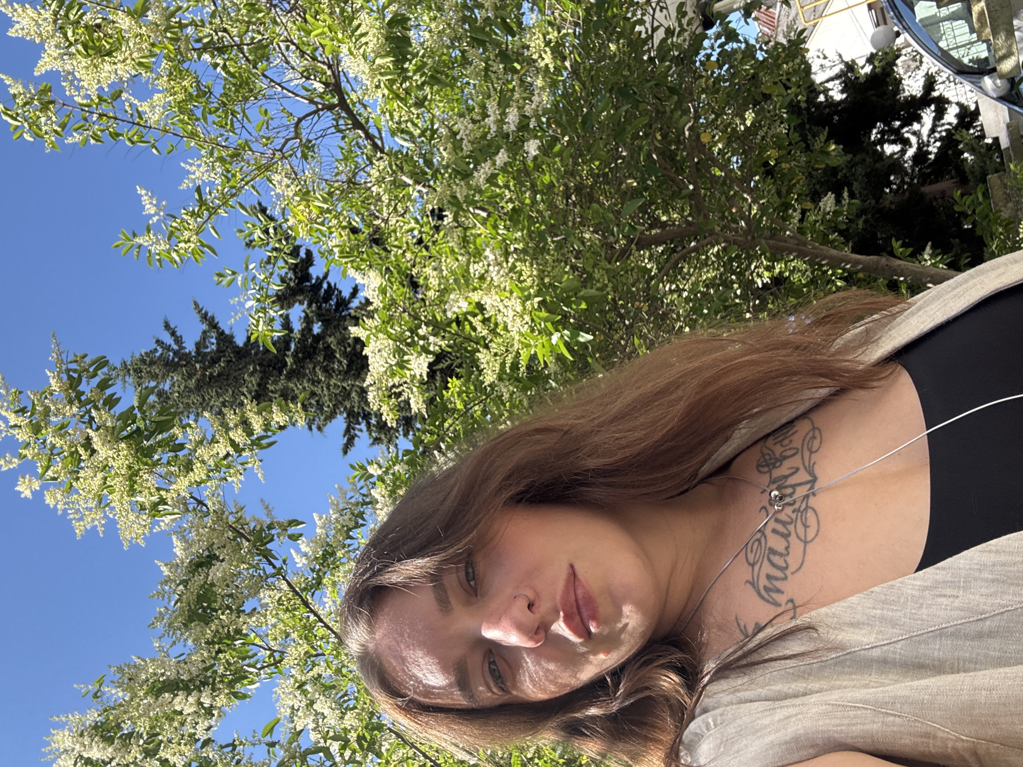 A female with long, wavy brown hair and a nose ring in her left nostril is positioned centrally, looking towards the camera. She wears a light-colored, sleeveless vest over a black top, revealing a large tattoo in Cyrillic script across her upper chest. The outdoor scene is brightly lit under a clear blue sky, indicating a sunny day in the city of Sochi, Russia. The background is dominated by lush green trees adorned with numerous clusters of small white flowers, suggesting springtime. A tall, dark coniferous tree stands on the right side of the image, while distant buildings and the partial view of a silver car are visible in the lower right corner.
