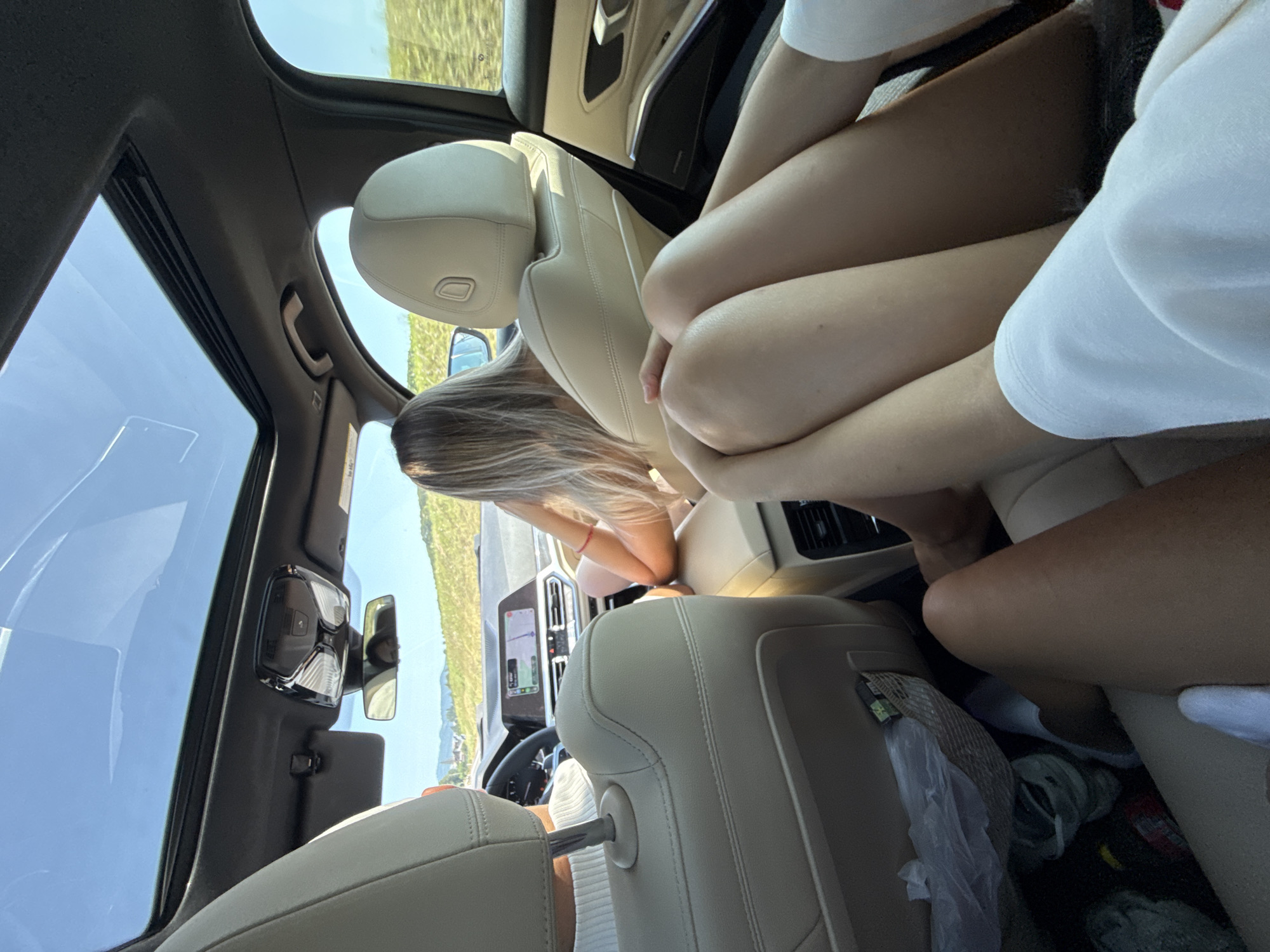 In the interior of a modern vehicle, two individuals are visible from behind. The person in the front passenger seat has blonde hair and is leaning back, resting their head on their hand. The driver, partially visible, is wearing a white sweater. In the foreground, the legs of another individual are prominent, appearing to be crossed. The car's interior features beige leather seats and a panoramic sunroof, through which a bright blue sky is visible. Outside the car, a rural landscape with rolling hills and vegetation under a clear sky can be seen. The scene suggests a journey through a scenic area, possibly in Dumbrăvița, Romania, given the context provided.