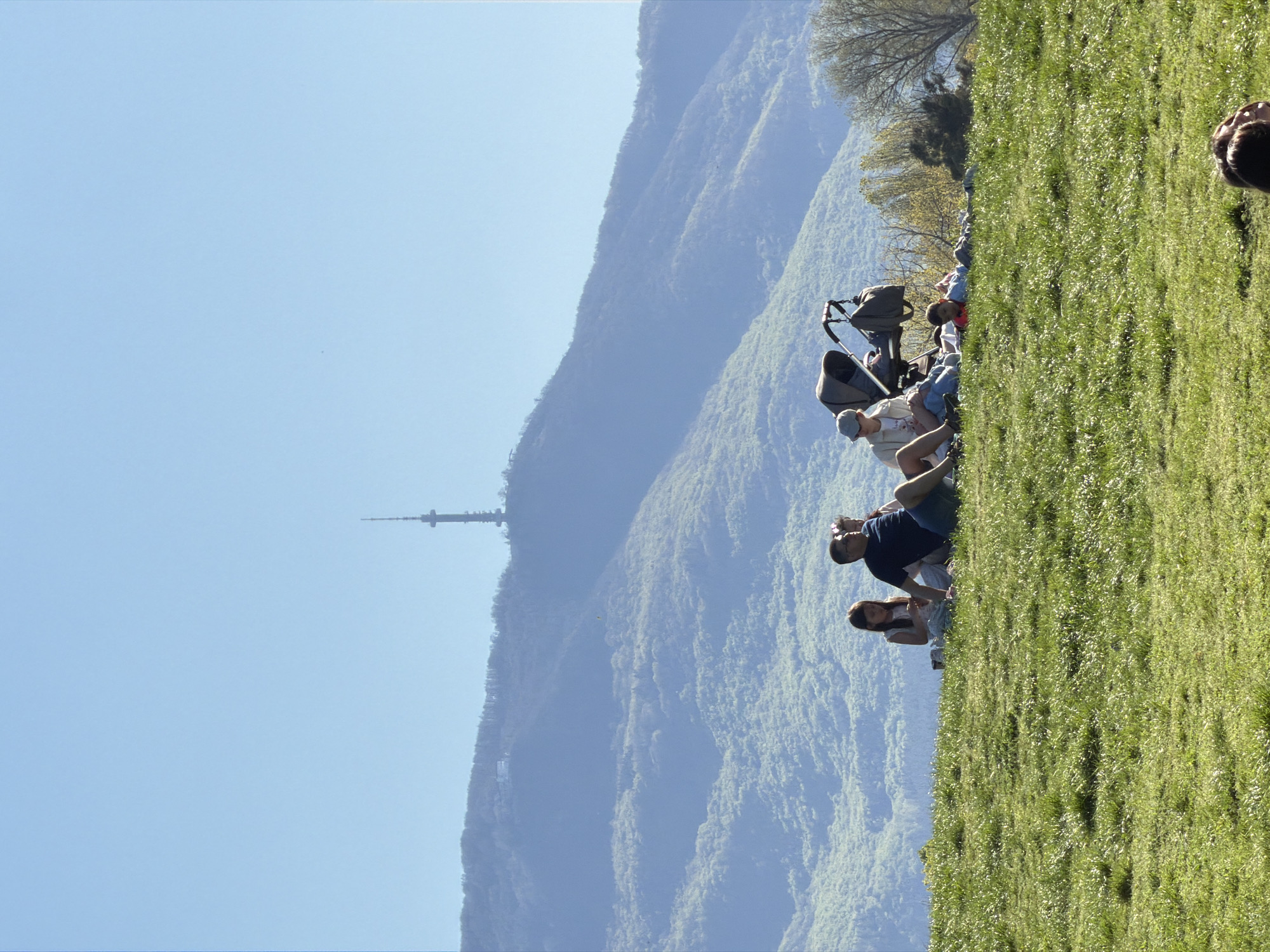 Here is a detailed description of the image:
The image shows a grassy hillside with people sitting and relaxing, with a mountain range in the background. The sky is clear and blue. A tall communications tower is visible on the mountainside.
A group of people of mixed ages is sitting on the grass. Some appear to be in conversation, while others are relaxing. There's a stroller parked among them.
The location is likely in or near Sofia, Bulgaria, given the presence of the Kopitoto TV tower on Vitosha Mountain.