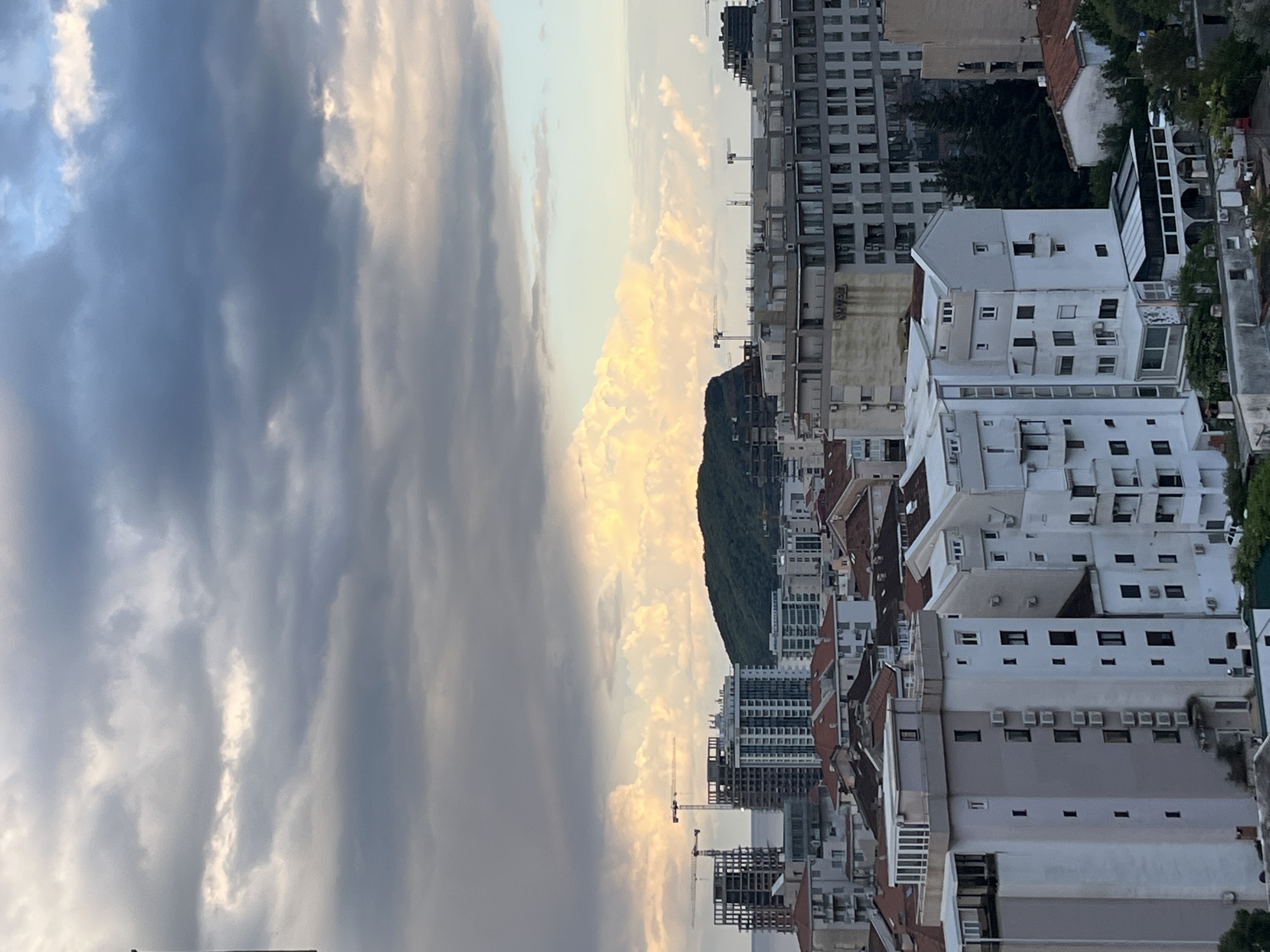 This image shows a cityscape in Budva, Montenegro, viewed from a high vantage point. The sky is filled with dark, striated clouds on the left, contrasting with lighter, golden-hued clouds towards the center. A tree-covered hill or small mountain can be seen among the buildings. In the foreground, there are numerous buildings, mostly white, with varying heights and architectural styles, some with red-tiled roofs. Construction sites with cranes are interspersed among the buildings. The perspective is tilted. There are no people visible in the frame.