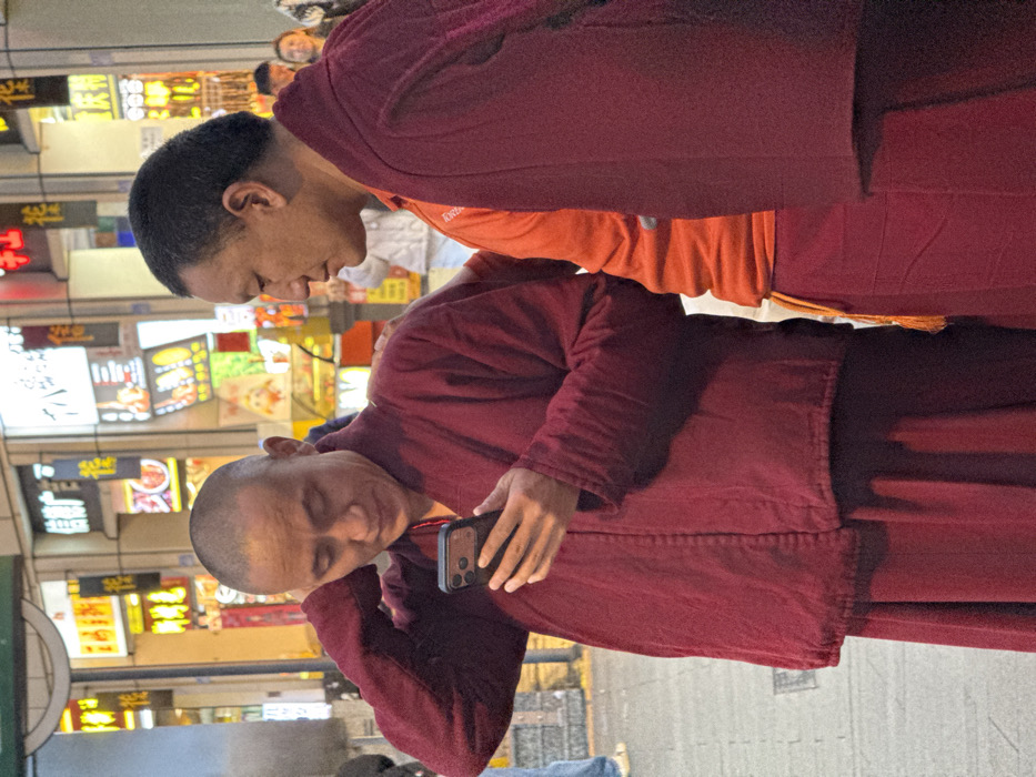 Two East Asian males, identified by their dark red monastic robes, stand on a paved urban street in Yuzhong District, China, at night. The male on the left, with a shaved head, holds a dark-colored smartphone with a multi-lens camera in his right hand, directing his gaze at its screen. His left arm is raised, with his hand resting near the back of his neck. The male on the right, with short dark hair, wears an orange shirt beneath his monastic robe, which is draped over his left shoulder. His right hand rests on the left monk's upper back, and he is looking towards the left. The background consists of a blurry but clearly visible commercial area, illuminated by numerous brightly lit storefront signs displaying Chinese characters.