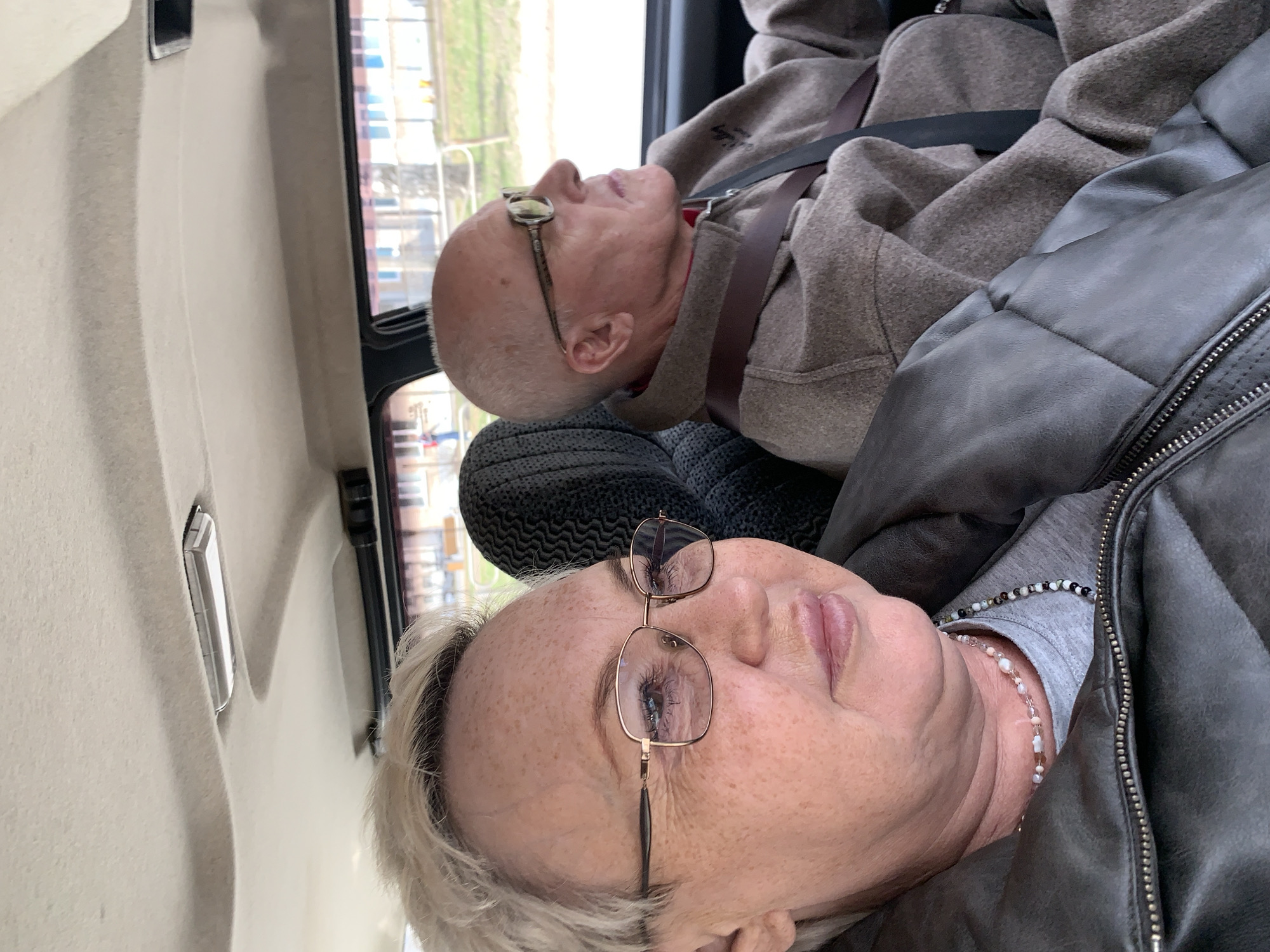 Two elderly people are seated in a vehicle. A woman with short, blonde hair and glasses, wearing a gray shirt and a black leather jacket, is in the foreground. Behind her is a man with a bald head and glasses, wearing a brown sweater and a seatbelt. They are both facing upwards. The background shows a blurred view through the window, possibly indicating they are in the city of Chaikovsky, Russia. The interior of the vehicle is light-colored, with a window frame visible. A headrest is visible behind the man.