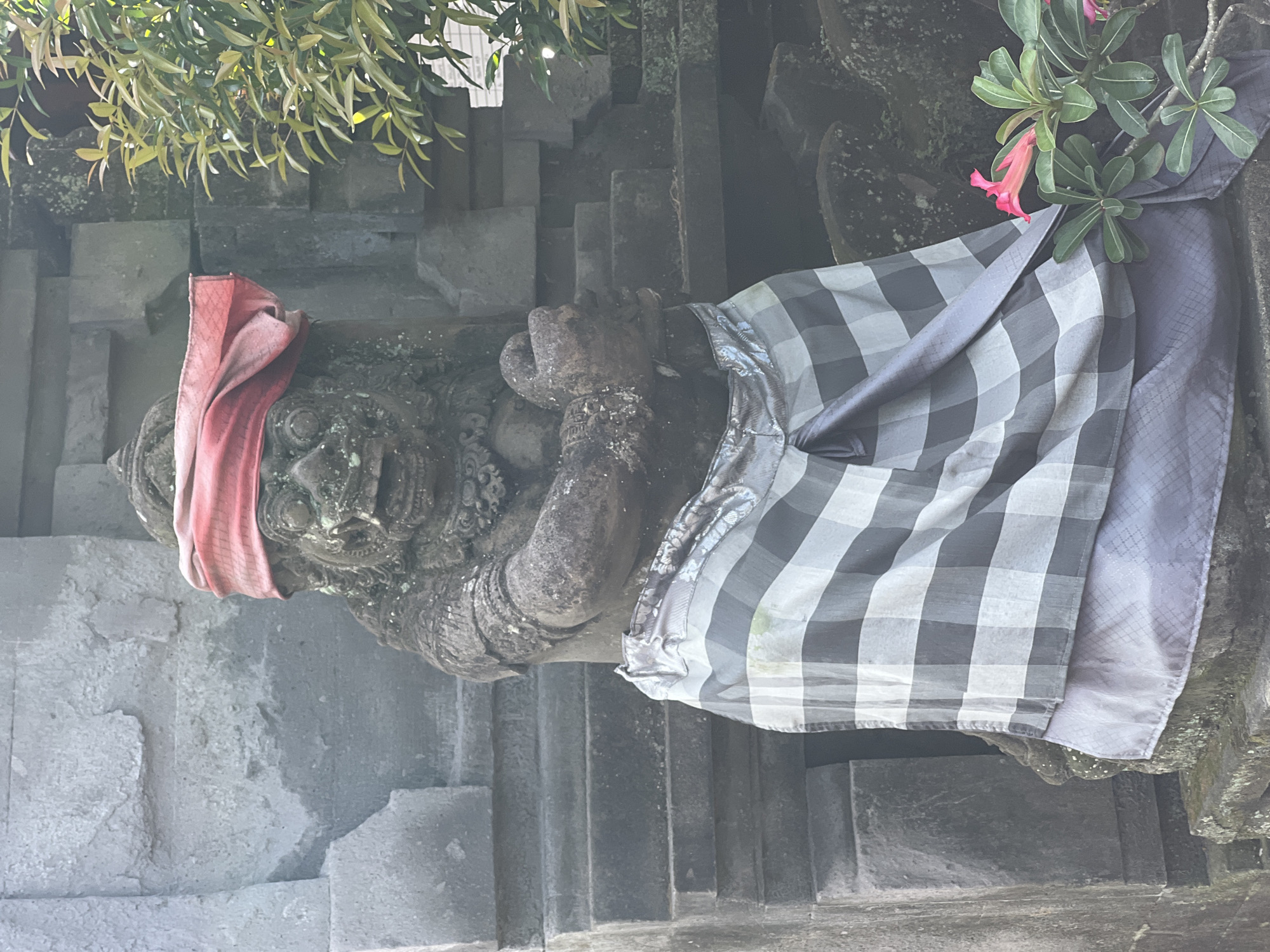 The image depicts a stone statue in Ubud District, Indonesia. The statue, possibly representing a deity or guardian, is adorned with a red cloth wrapped around its head and a black and white checkered textile draped across its body. The statue is situated in a stone structure, likely part of a temple or religious site. A plant with green leaves and a single pink flower grows adjacent to the statue. The environment appears humid, with stone surfaces showing signs of weathering and possible moss growth. The composition is slightly tilted.