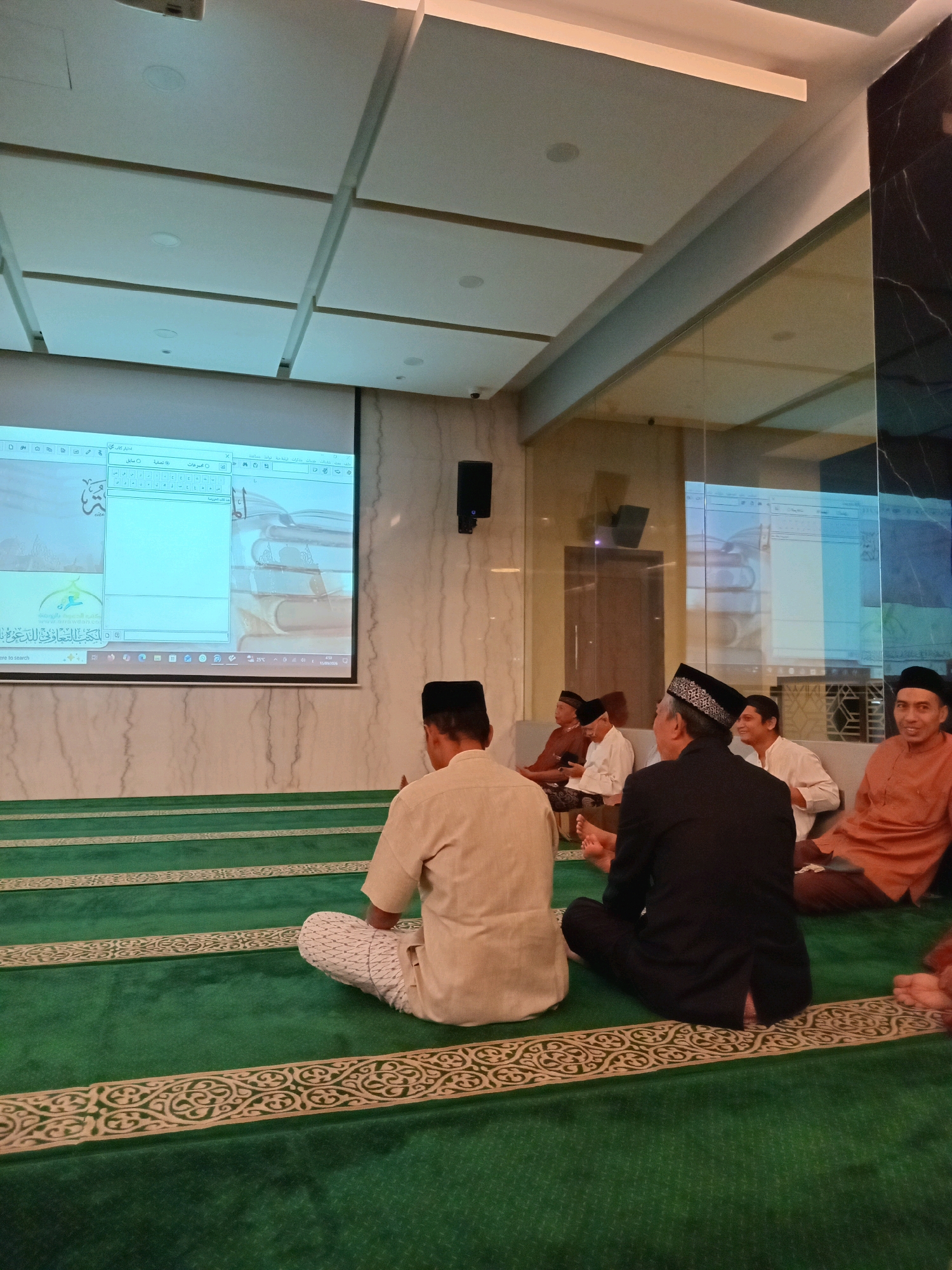 In a prayer hall, likely in Randuagung, Indonesia, several men are seated on a green carpet with patterned borders. A large projection screen displays a computer interface with Arabic text, possibly a presentation or software demonstration. The men are dressed in traditional Islamic attire, including peci caps and long shirts. One man in the foreground faces away from the camera, while others are visible in the middle ground and background, some engaged in conversation. The hall has modern lighting and a mirrored wall, suggesting a contemporary setting.