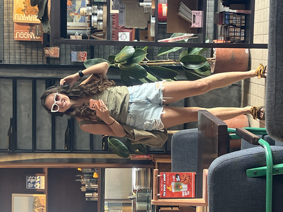 A woman with long, curly brown hair and sunglasses poses indoors. She's wearing a light green shirt and distressed denim shorts. She holds a cell phone in her hand. Shelves with various items and books are in the background. The woman appears to be in a café or shop, as indicated by furniture and decorative elements nearby.