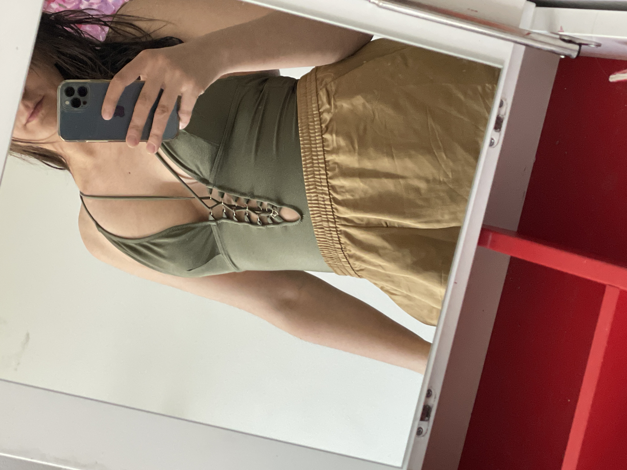 A woman is shown from the chest down in a mirror selfie. She is wearing an olive green tank top with crisscross detailing on the chest, paired with gold shorts. Her phone is visible in her hand, obscuring part of her face. The setting appears to be indoors, with a white vanity and a red interior partially visible in the reflection. The city and location are unknown.