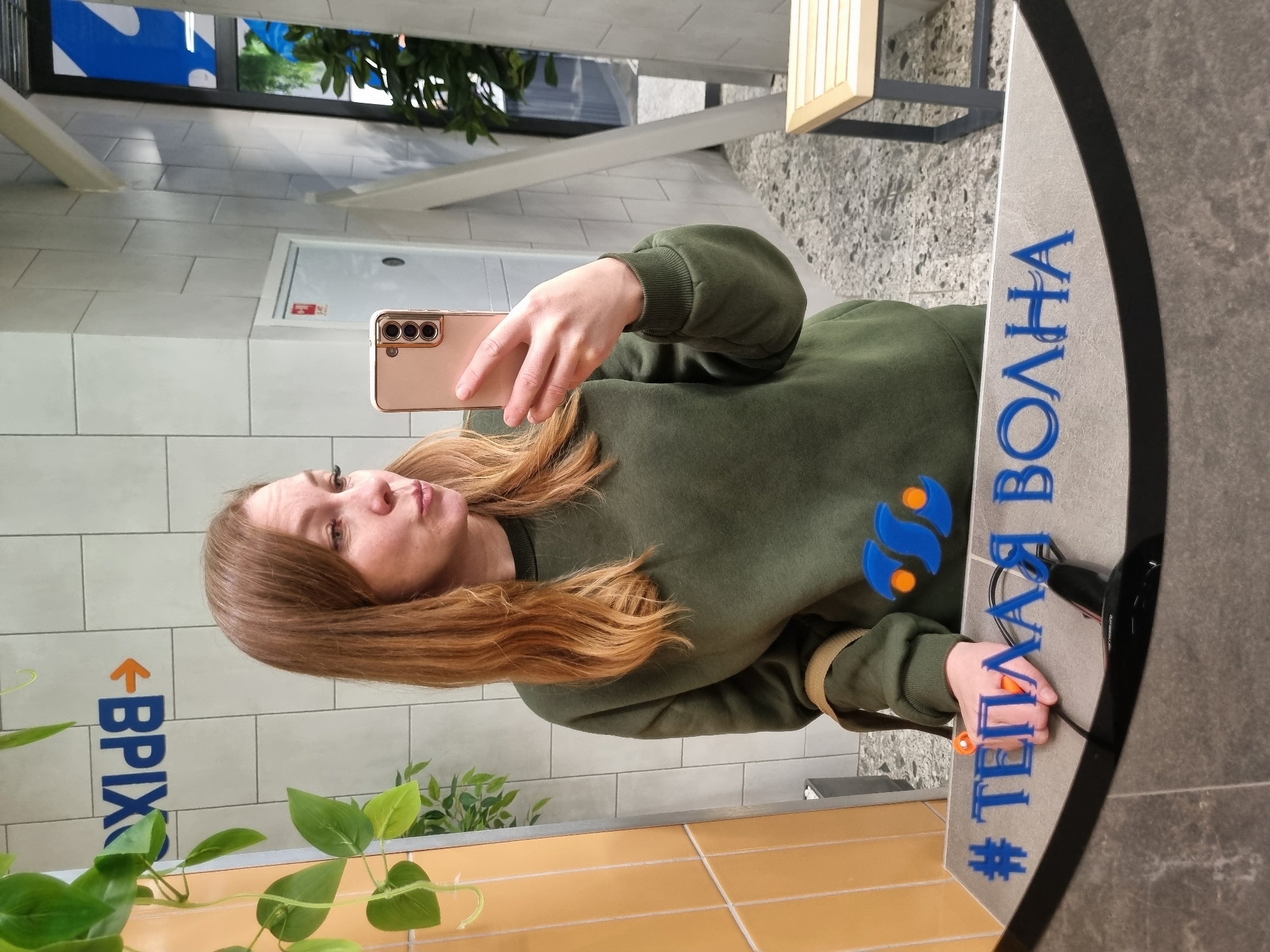 A person with brown hair is taking a selfie in a restroom. They are holding a light pink smartphone with three camera lenses on the back. The person is wearing an olive green sweatshirt. The restroom has light grey tiling, and a yellow tiled countertop is visible in the lower portion of the frame. A potted green plant sits on the countertop. Blue text is displayed in the reflection of the mirror, with the word "Волна" visible, along with a blue logo comprised of two water droplet-like shapes. The logo appears to be for "Теплая Волна" based on the hashtag preceding the text. Also in the reflection, the acronym "ВРІХ" is displayed in blue vertically on the wall. The location is Votkinsk, Russia.