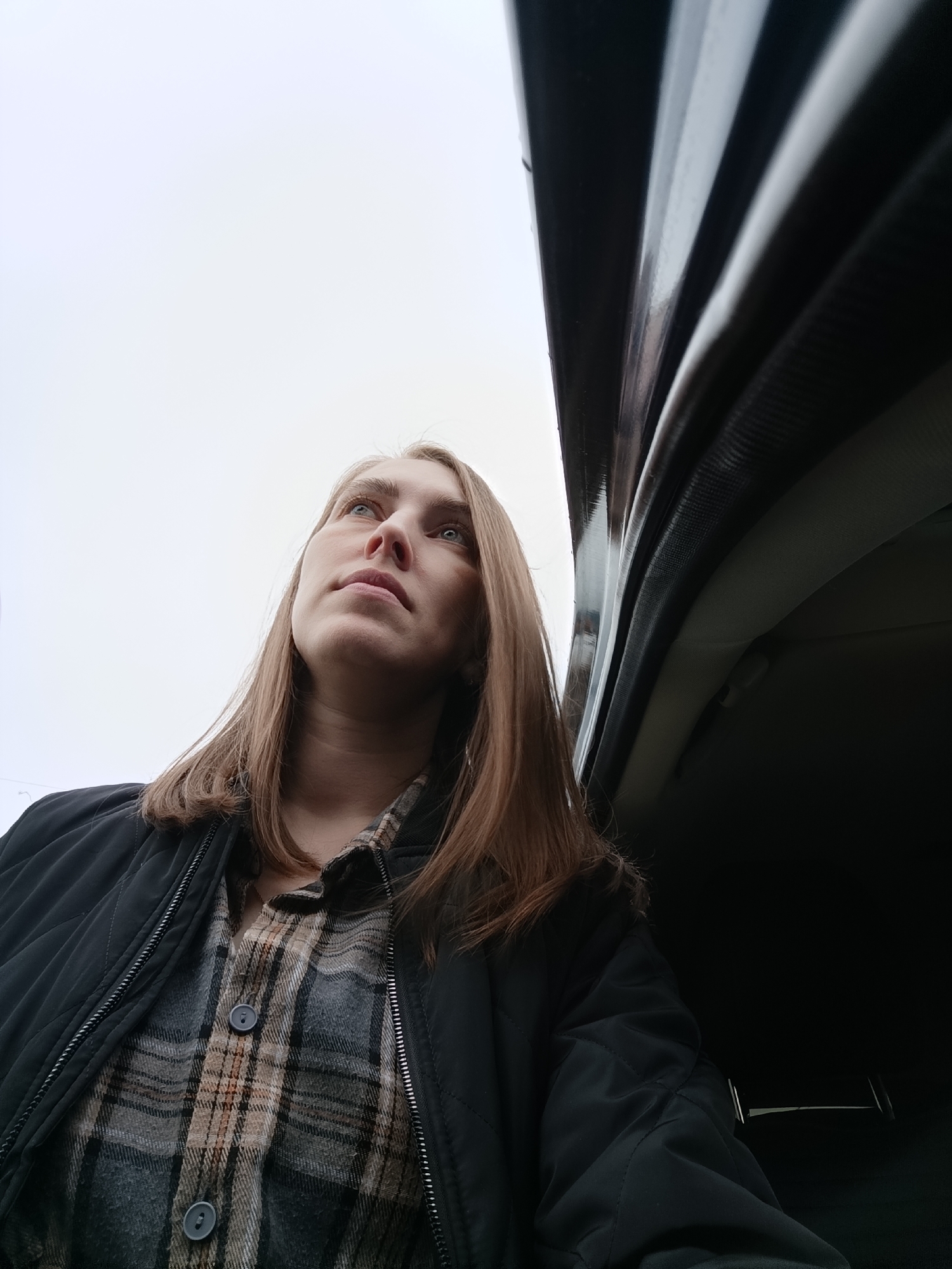 A low-angle medium shot captures a fair-skinned woman with light brown hair looking upwards. She is wearing a dark-colored quilted jacket over a buttoned plaid shirt. The background consists of an overcast sky and the interior of a vehicle, possibly a car, with dark trim and upholstery. The woman's location and whether the photograph was taken in Chaikovsky, Russia, cannot be determined from the image.
