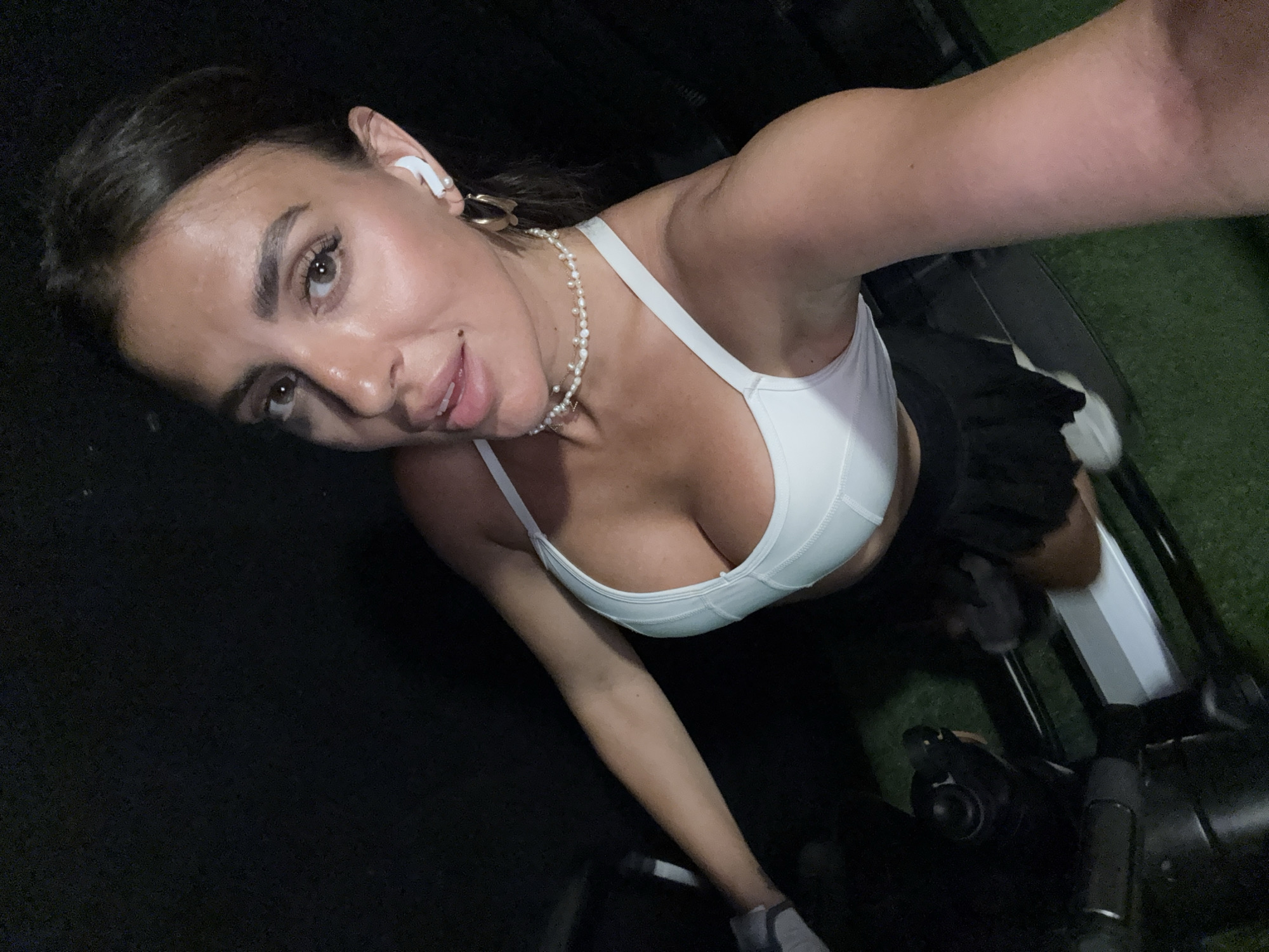 A selfie of a woman in workout attire in a dark indoor location, presumably a gym. She is wearing a white sports bra and pearl necklace. She has dark hair pulled back, is wearing an earbud in one ear, and has on a gold earring. She is wearing gloves. The lower portion of the image shows the handlebars and equipment of an exercise bike. The background is dark and indistinct. It is impossible to confirm the location is in North Kuta, Indonesia, without additional information.