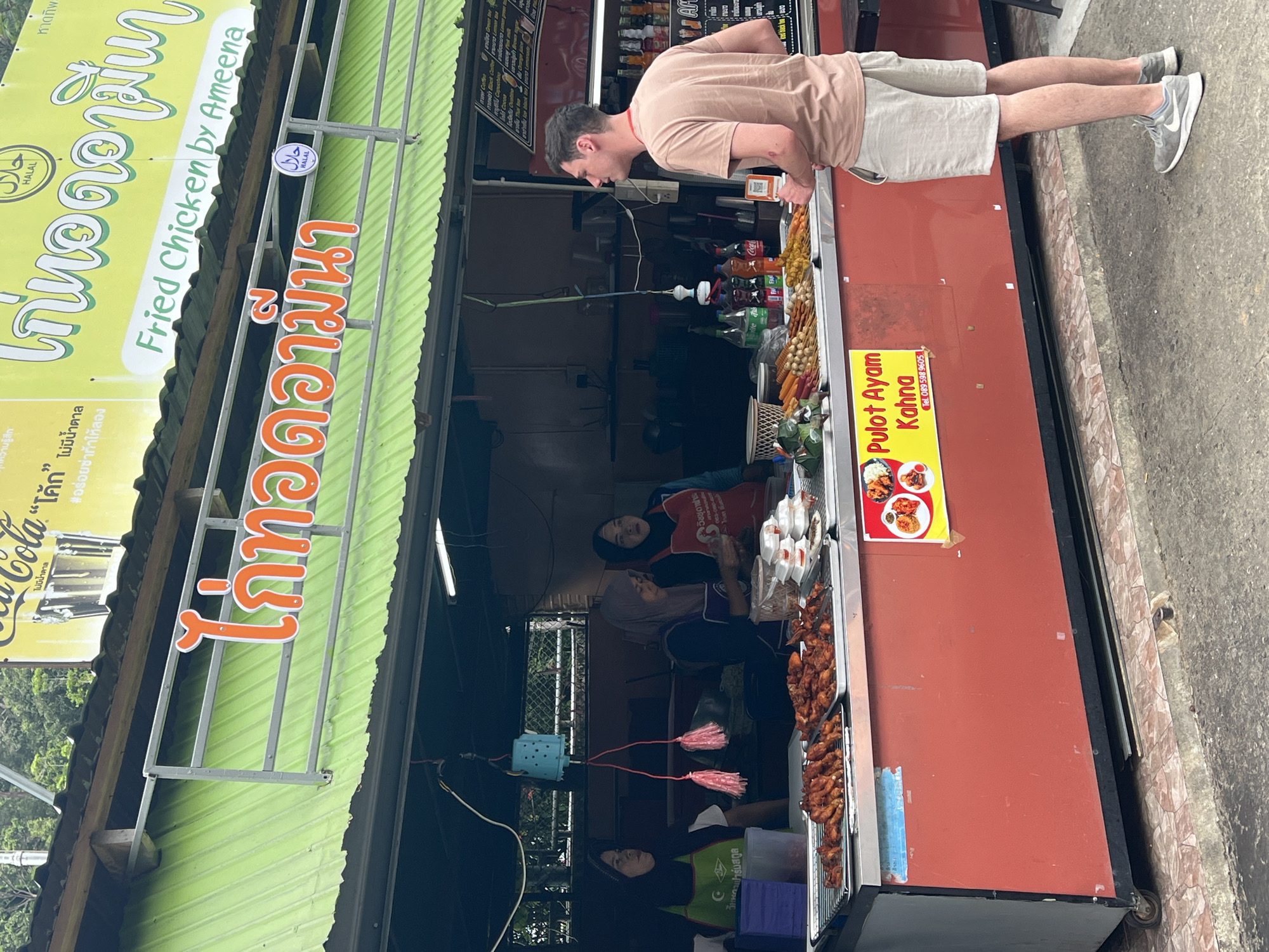 This media file captures a vibrant street food scene in Tambon Wang Prachan, Thailand. The primary subjects are people and the food stall they are interacting with. A man, wearing a light beige t-shirt and light-colored shorts, is leaning over the counter, appearing to order or observe the food. He is dressed casually, suggesting a warm day.
The stall itself is the central object, with a bright green corrugated metal roof and a prominent red facade. A large yellow sign with Thai script and the words "Fried Chicken by Ameena" in English, along with "Pulot Ayam Kahna," indicates the type of food sold. The sign also features the Halal logo, suggesting the food is prepared according to Islamic dietary laws. Below the main signage, a Coca-Cola advertisement is visible, partially obscured.
The scene is busy with activity behind the counter. At least three women, two of whom are wearing hijabs and aprons, are working at the stall. They appear to be preparing or serving food, with trays of fried chicken and skewers prominently displayed. The atmosphere seems bustling and transactional, typical of a popular street food vendor.
Notable details include the various food items, such as skewers of what appears to be fried chicken or meat, displayed in trays. Several bottles of drinks, including Coca-Cola, are visible on shelves behind the counter. The presence of the Halal logo and the mix of Thai and Malay text ("Pulot Ayam Kahna") suggest a diverse cultural context. The ground beneath the stall is paved, indicating an urban or semi-urban setting. The lighting suggests it is daytime, though the exact time of day is not clear. The overall impression is a lively and authentic street food experience in Thailand.