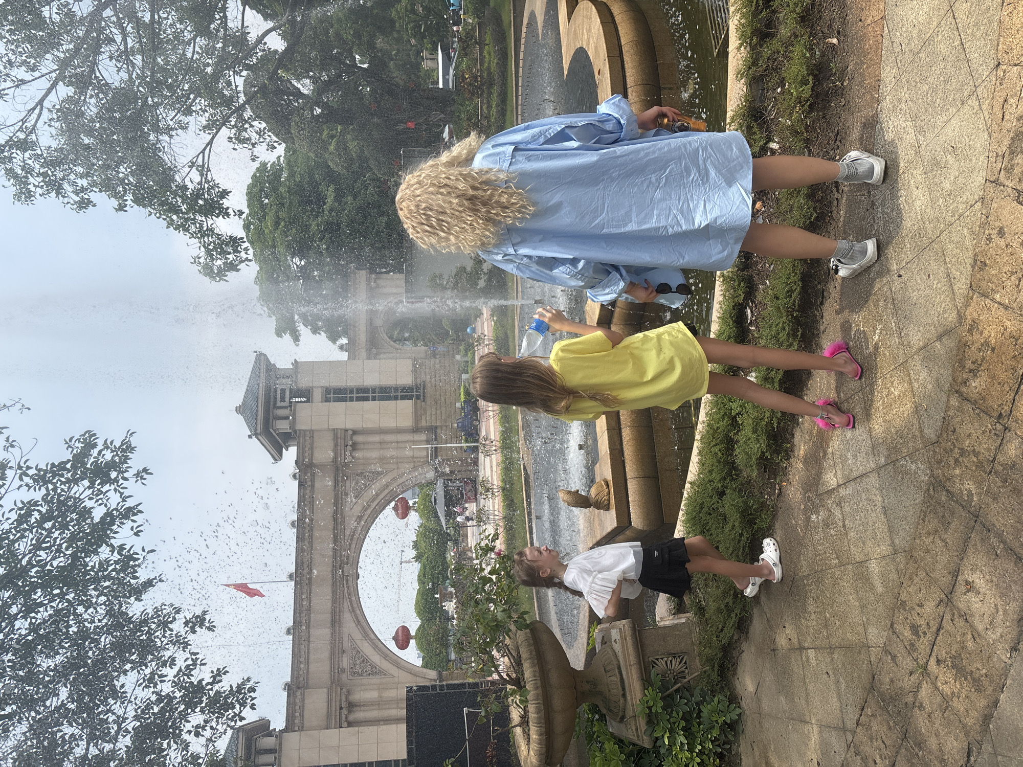 In Nanhai District, China, three individuals stand near a fountain in an outdoor setting. A woman with blonde curly hair, wearing a light blue oversized shirt and grey socks with white sneakers, holds a can and a pair of sunglasses. To her left, a young girl in a yellow t-shirt and pink slides stands facing away from the camera, holding a clear plastic water bottle. To the left of the girl in yellow, another young girl in a white ruffled shirt and a black skirt, wearing white clogs, faces left with her hands on her hips. A large stone fountain with water jets is visible in front of them. In the background, an arched stone structure with decorative carvings stands under a cloudy sky. Red lanterns are hung on either side of the archway. Trees and greenery surround the area.