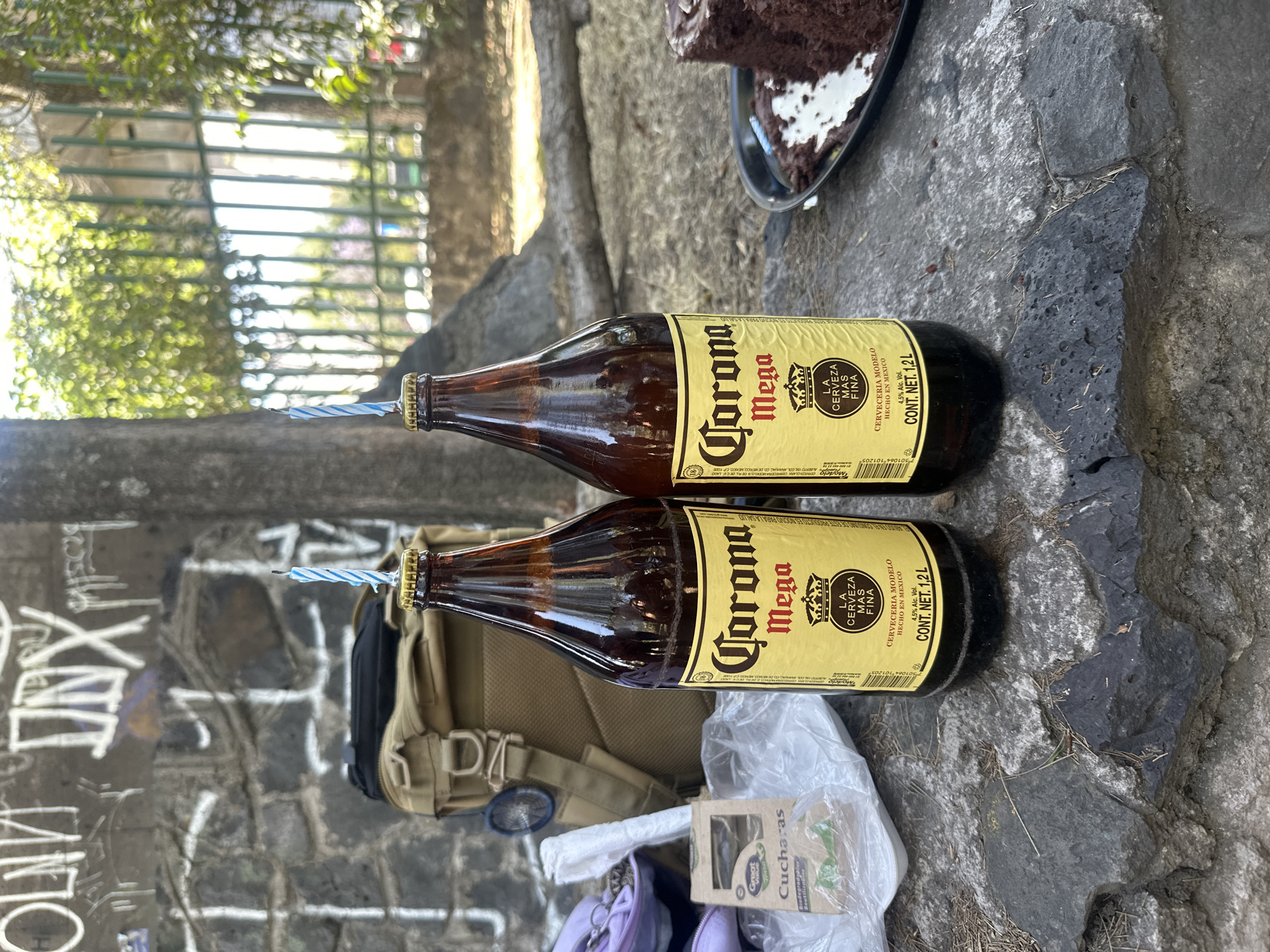 The image depicts an informal outdoor celebration in Mexico City, Mexico. Two large, brown glass bottles of "Corona Mega" beer are centered, each featuring a blue and white striped birthday candle inserted into its neck. A slice of chocolate cake with white frosting on a dark plate is partially visible in the upper right corner, further indicating a birthday event.
The items are arranged on a dark, rough stone surface, possibly a low wall or the ground. Behind the lower beer bottle, a tan-colored backpack is visible. Next to it, a white plastic bag holds a small cardboard box labeled "Great Value Cucharas," indicating biodegradable and reusable spoons. A purple item is also partially seen near the bag.
The background features a dark stone wall covered with various white graffiti tags. Beyond the wall, green foliage and trees are visible, with a metal fence or structure in the distant, out-of-focus background. The scene is illuminated by bright natural light, suggesting a sunny daytime setting.
Visible text on the beer labels includes "Corona Mega," "LA CERVEZA MAS FINA" (The Finest Beer), "CERVECERIA MODELO," "HECHO EN MEXICO" (Made in Mexico), "4.5% Alc. Vol.," and "CONT. NET. 1.2L," indicating the large volume of the bottles. The text "NO ES NOCIVO PARA LA SALUD" (It is not harmful to health) is partially visible. The box of spoons is labeled "Biodegradable Reutiliz." The composition suggests a casual and festive outdoor gathering.