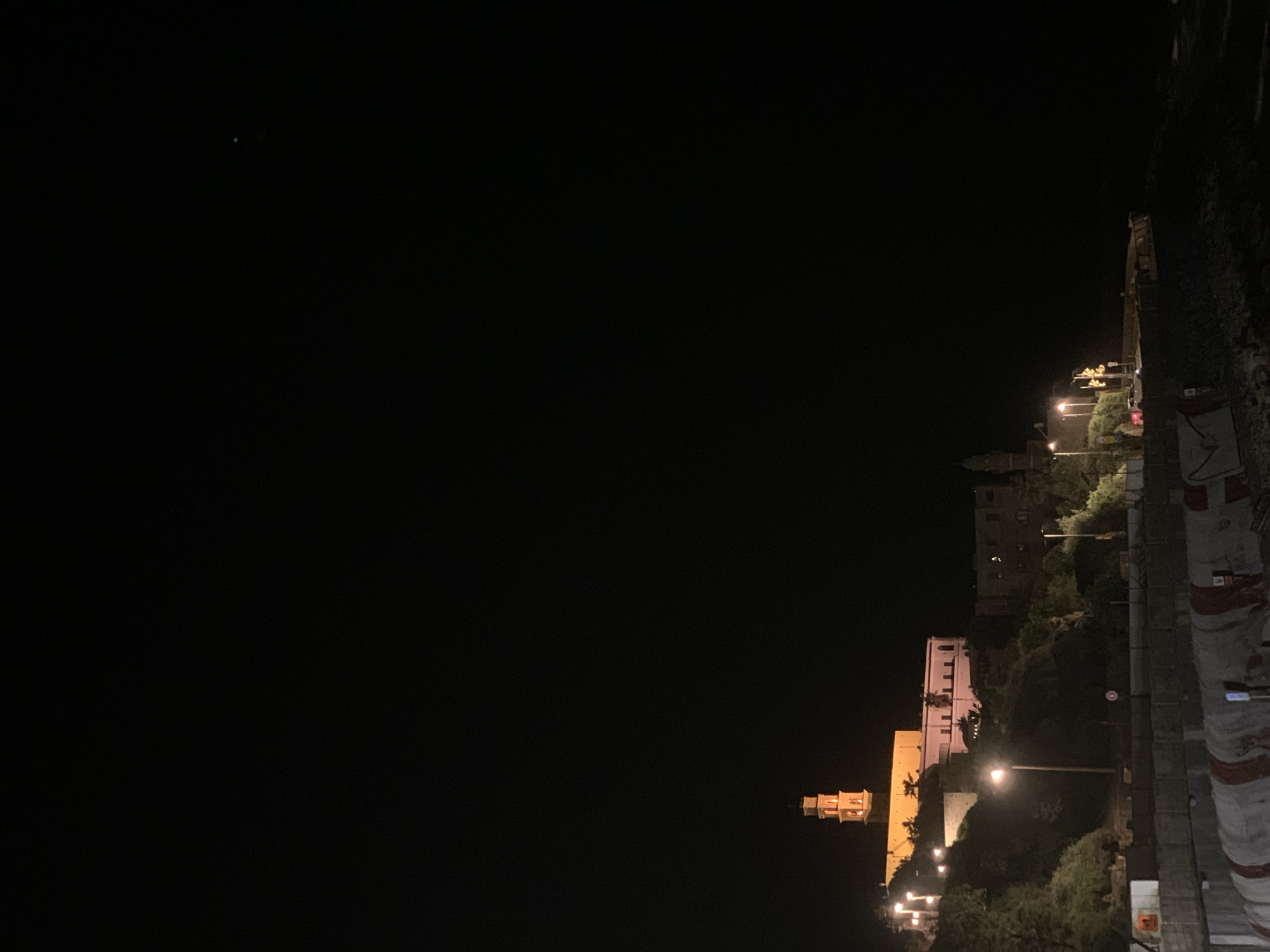 The image depicts a night scene in the old city of Jaffa, Tel Aviv, Israel. The frame captures the illuminated buildings along the hillside overlooking the Mediterranean Sea. The street level shows pedestrian walkways, bordering the building facades on the hill. Streetlights provide some illumination, with the buildings themselves emanating a soft, warm glow. The sky is mostly black, with a faint star visible.