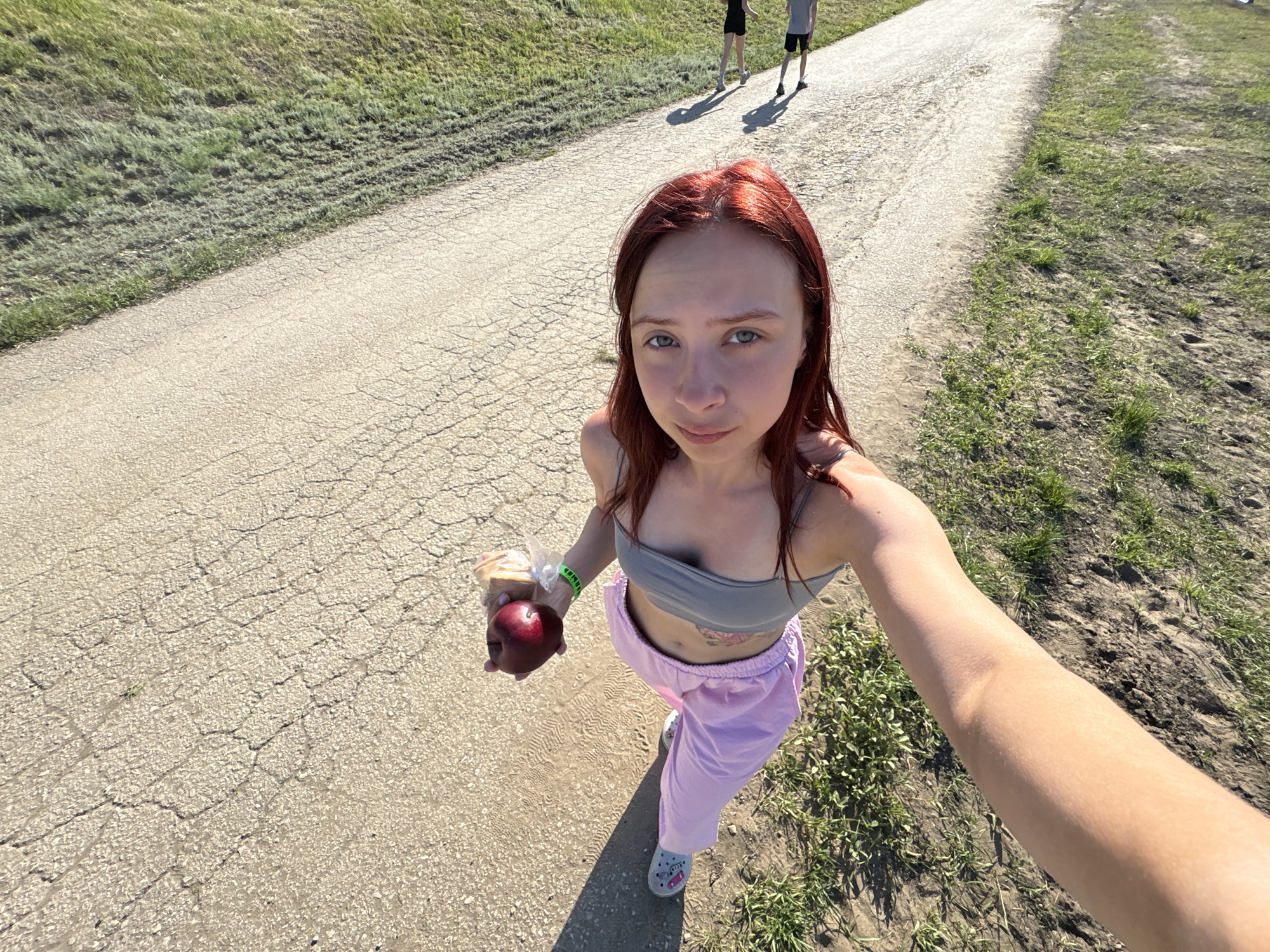 A young, red-haired woman stands on a cracked concrete path, holding a red apple and a plastic-wrapped item. She is wearing a gray tank top and lavender sweatpants, and Crocs. The scene is set outdoors, likely a park or recreational area, with grass bordering the path on either side. In the background, two indistinct figures are walking away from the camera on the same path. The woman is likely taking a selfie, evident by the outstretched arm. The location is outdoors in the vicinity of Khvalynsk, Russia.