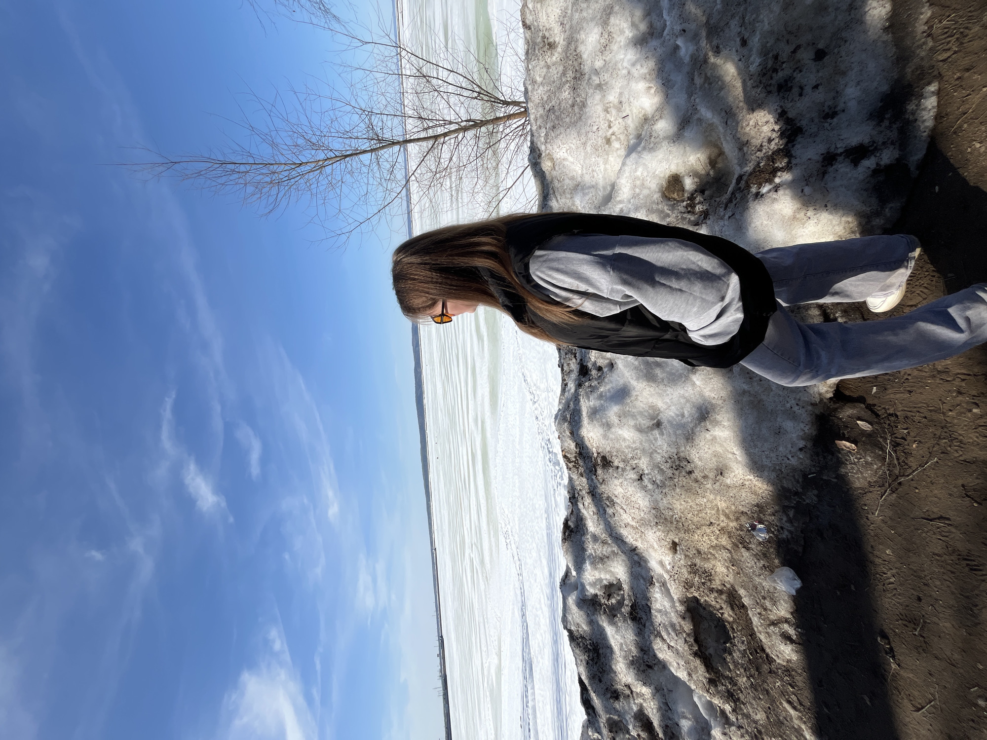 A female individual, viewed from the right side, stands on a dirt path alongside a bank of melting snow mixed with earth. She has long brown hair and wears orange-tinted sunglasses, a black puffy vest over a grey long-sleeved shirt, and light grey wide-leg pants with white sneakers. Her hands appear to be in her pockets or obscured. She is looking left, towards a vast, partially frozen body of water, identified as the Votkinsk Reservoir in Chaikovsky, Russia. The ice on the reservoir is predominantly white, with areas of greenish slush or open water, and shows horizontal markings. On the distant horizon across the water, industrial structures including cranes, buildings, and power poles are visible. A leafless deciduous tree with thin branches stands to the right of the individual on the snowy bank. The sky above is clear blue with sparse, wispy white clouds. The ground in the immediate foreground shows patches of dark earth and snow, with some visible litter, including a small clear plastic bag and a white wrapper. Shadows are cast towards the lower right. The scene depicts an outdoor environment during late winter or early spring.