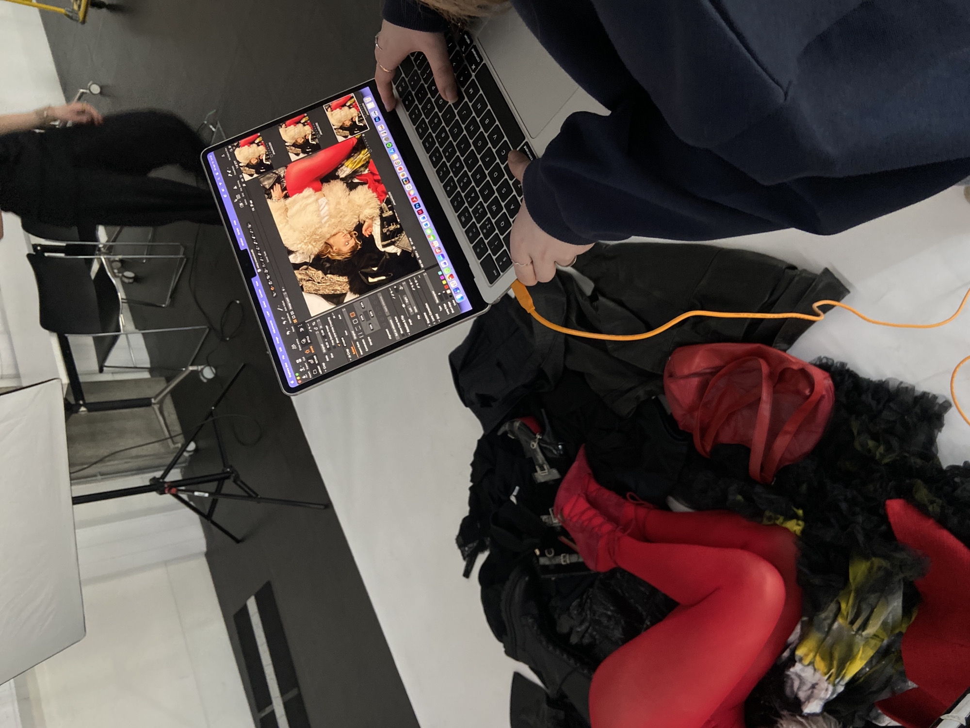 The image captures a scene within a photography studio, likely in London, United Kingdom. A laptop displays a series of images, with what appears to be raw footage of a model in costume with bright red tights and shoes. The individual manipulating the laptop, wearing a dark blue sweater, is shown engaging with the keyboard. The camera is likely tethered to the laptop with the visible orange cable. The setting includes photographic equipment such as a light stand, as well as props or wardrobe elements scattered on a white surface. A person in dark clothing is partially visible near a chair. The environment has a stark contrast between white and black.