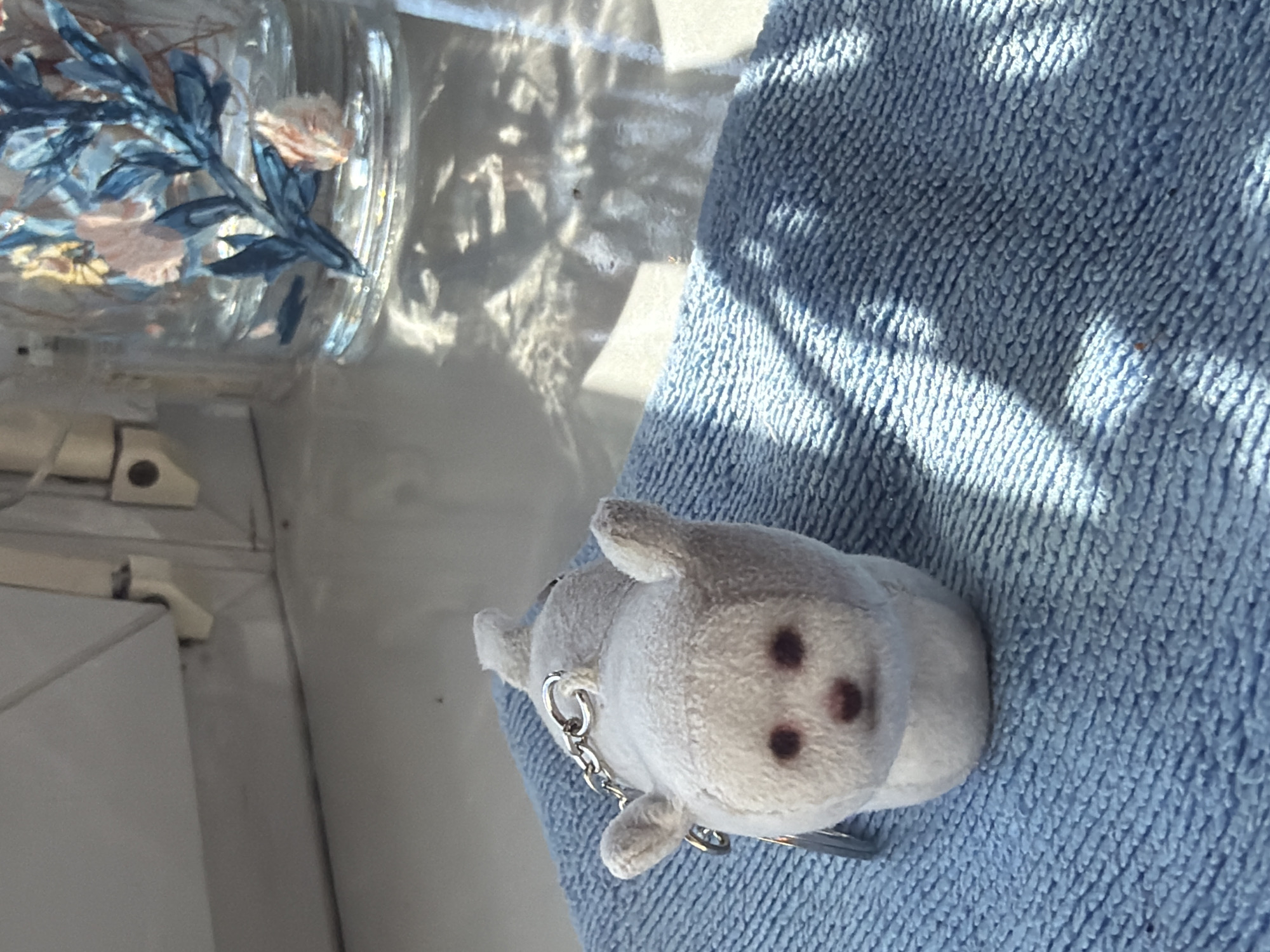 The image shows a close-up of a small, gray, plush toy animal with dark eyes and a chain attached, lying on a blue, textured surface. The toy appears to be a keychain or a decorative charm. In the background, to the left, is a clear glass vase containing artificial floral decorations with blue and pink elements. Sunlight casts dappled shadows on both the toy and the blue surface, suggesting it is daytime. The overall setting is unclear, but the presence of the vase and the way the light falls hints at an indoor environment, possibly near a window. There is no visible text in the image.