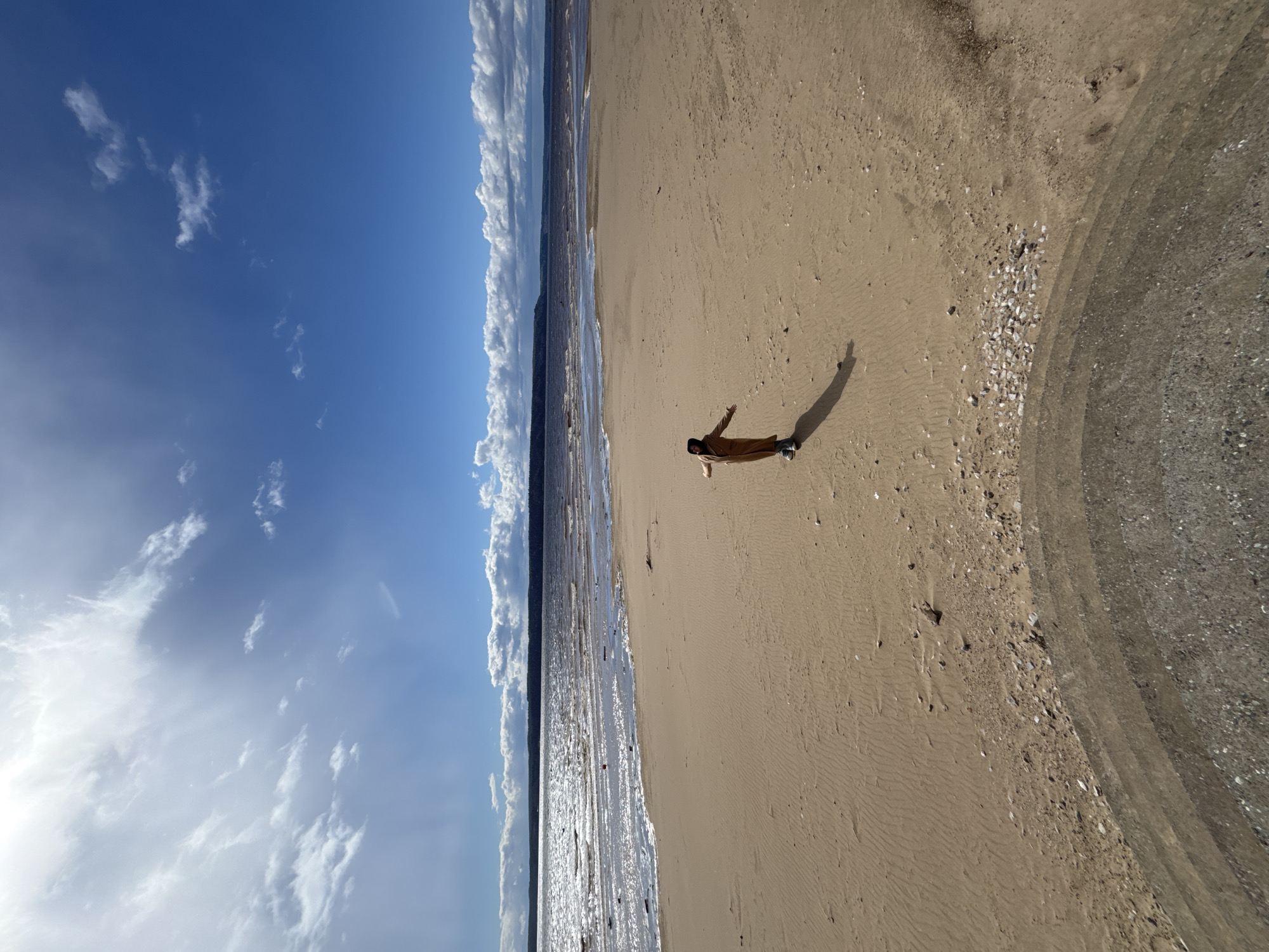 The vertically oriented image captures an expansive outdoor scene, featuring a sandy beach bordering a calm body of water, likely the Kama Reservoir near Polazna, Russia. Above, a bright blue sky is adorned with scattered white clouds.
A single individual is the central subject, seen lying or reclining on the golden sand with their arms slightly spread. They are dressed in a light brown coat and dark clothing. A distinct, elongated shadow stretches behind the person on the textured sand, indicating the sun is relatively low in the sky, suggesting either early morning or late afternoon.
The sandy shoreline displays subtle ripples and scattered small stones and shells where it meets the water. The water itself appears calm, reflecting the bright sky, with gentle waves breaking near the shore. In the distance, across the water, a dark, low-lying landmass, possibly forested hills, is visible beneath a band of white cumulus clouds that stretch across the horizon. The sky transitions from a lighter blue near the horizon to a deeper hue overhead, dotted with wispy white clouds. The overall atmosphere evokes a sense of peace, relaxation, and solitude. There is no visible text in the image.
