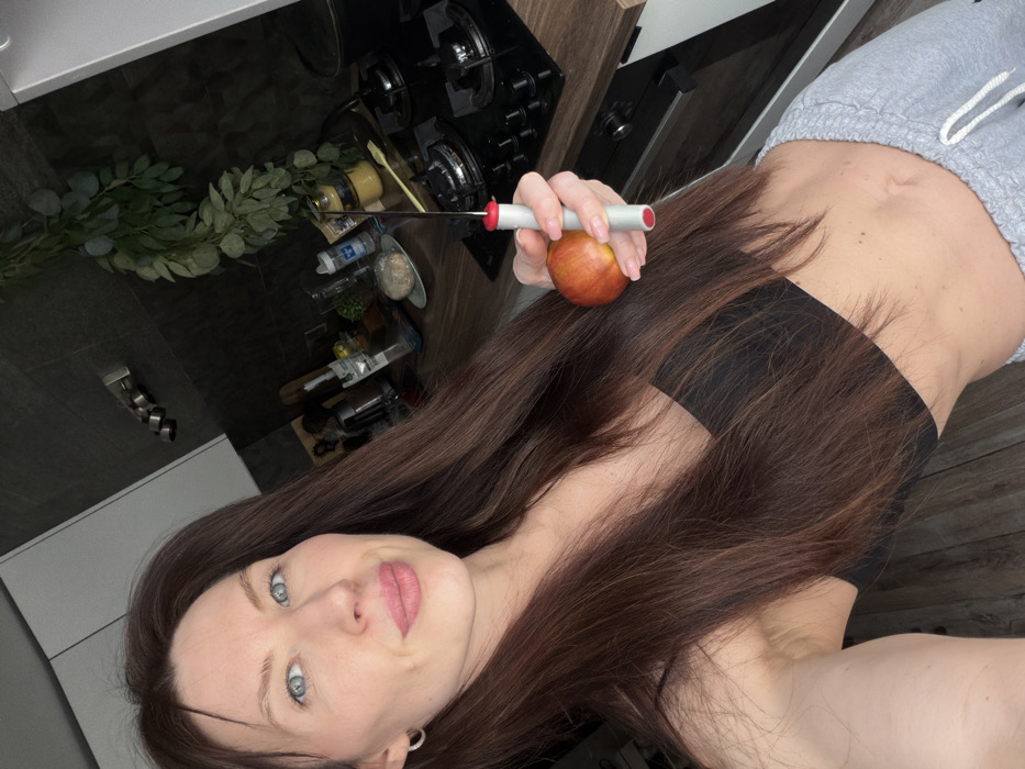 A female subject is depicted indoors in a kitchen environment, identified as being in Perm, Russia. The subject, an adult female with long, straight, dark brown hair and light blue eyes, is taking a selfie, looking directly at the camera with a slight smile. She is wearing a black bandeau-style top and grey sweatpants with a visible navel. Her left ear displays a small silver hoop earring. In her left hand, she holds a red and yellow apple. Her right hand grasps a grey and red fruit corer, with its metal spike positioned above the apple. The background reveals a modern kitchen featuring a dark grey tiled backsplash and a wooden-patterned countertop. A black gas hob with four burners and control knobs is visible on the countertop, with a black oven door partially visible beneath it. White overhead cabinets are situated on the left, and a chrome-colored fixture with multiple bars is mounted on the dark tiled wall. Various kitchen items, including bottles, jars, and a white plate with a light-colored, round food item, are arranged on the countertop. A cluster of green leafy decor hangs against the dark tiled wall. The flooring consists of light-colored wood or laminate.