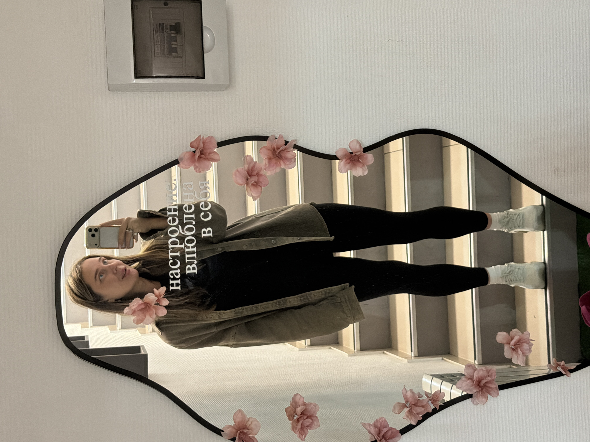 A full-body selfie of a woman is reflected in a decorative mirror affixed to a white wall. The woman is holding a smartphone in her left hand, capturing her reflection. She is wearing black leggings, a black top, an unbuttoned green shirt, and white socks. The mirror is irregularly shaped with a black frame and is adorned with pink flower decorations. The reflection in the mirror shows a staircase with light-colored treads and white risers. A white electrical panel is mounted on the wall above the mirror. Inscribed text is visible on the mirror's surface. The scene is located in Chaikovsky, Russia, as indicated by the text on the mirror, "настроение. влюблена в себя," which translates to "mood. in love with myself."