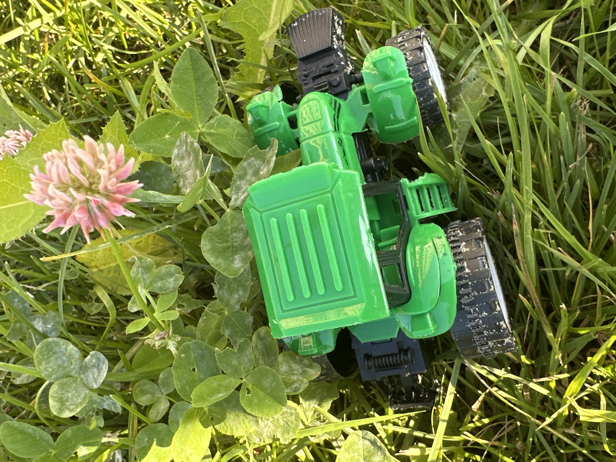 A green plastic toy tractor is lying on its side in thick green grass and clover, with a pink clover flower visible in the upper right portion of the frame. The tractor is oriented with its back to the viewer. The grass is illuminated by bright sunlight, casting shadows. The scene appears to be outdoors, possibly in a park or backyard. There is no discernible information about the city of Znamya Oktyabrya, Russia, in the image.