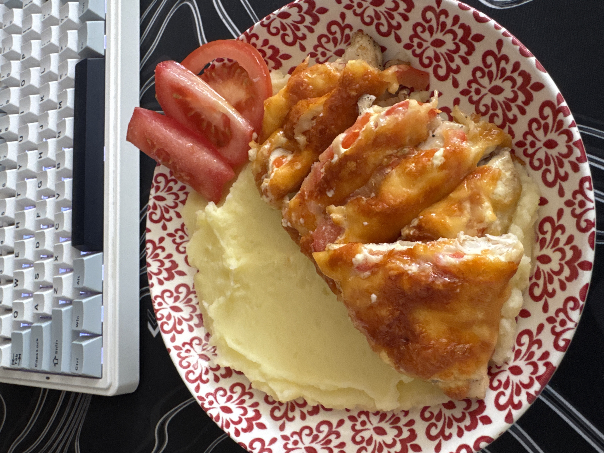 A high-angle shot captures a plate of food set against a black patterned surface, with a white keyboard partially visible on the left. The plate, decorated with a red floral pattern, contains mashed potatoes, sliced tomatoes, and a serving of what appears to be baked or broiled chicken topped with melted cheese and tomato. The lighting is bright, illuminating the food and keyboard. The photograph also has a keyboard located to the left of the plate. The location is likely Perm, Russia, as indicated in the prompts.