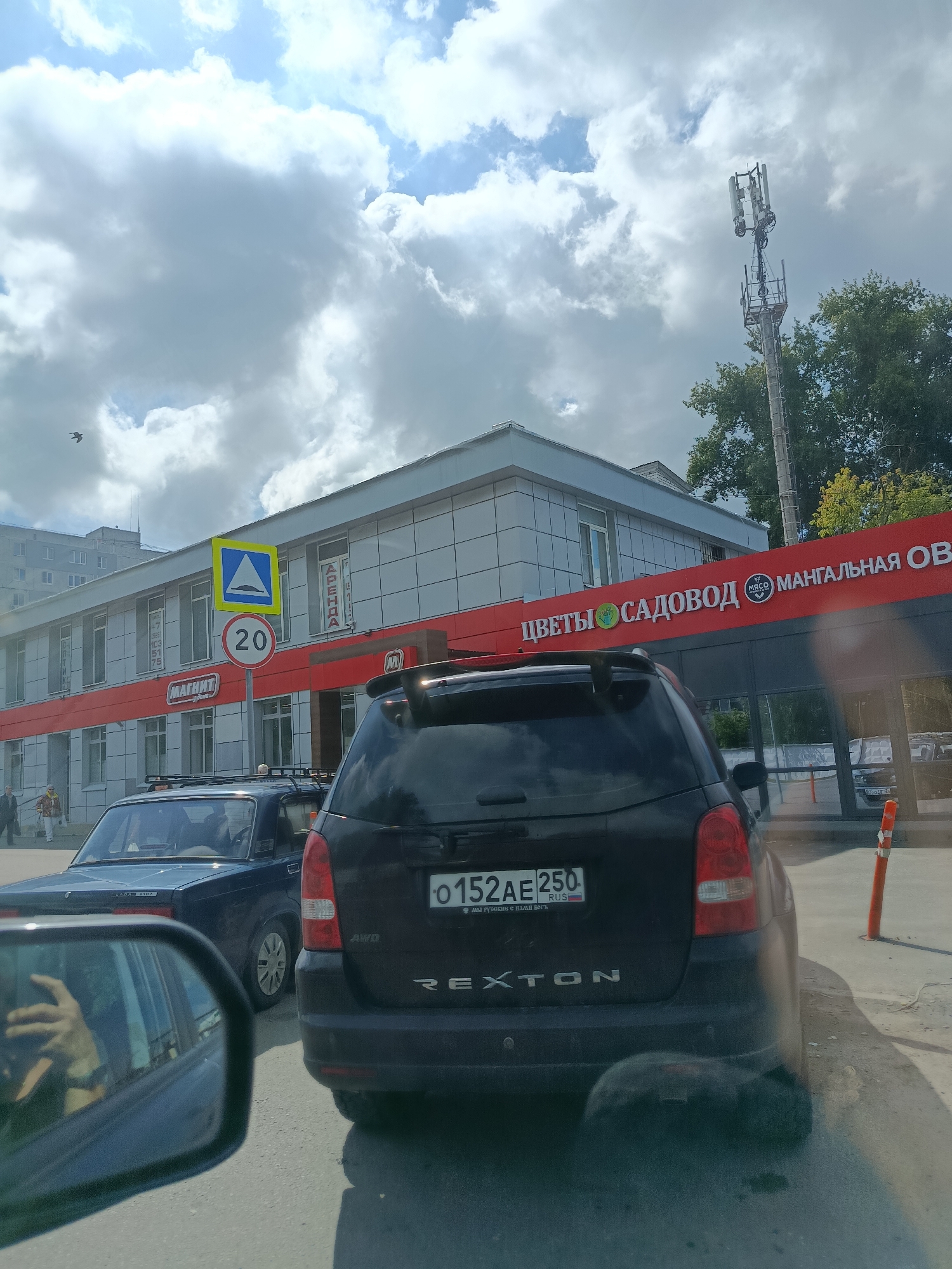A black SsangYong Rexton SUV is parked on a paved road, possibly a street or a parking lot, in Orekhovo-Zuyevo, Russia. The SUV has a license plate that reads "o 152 AE 250 RUS". In front of the Rexton, a dark blue sedan is partially visible. To the left, a reflection of a person holding a smartphone is seen in the driver's side mirror of the Rexton. Behind the Rexton is a building with a red awning that has white lettering, some of which reads "ЦВЕТЫ САДОВОД МАНГАЛЬНАЯ ОВ". The building also has "МАГНИТ" signage. A speed limit sign indicating "20" and a pedestrian crossing sign are visible on poles in front of the building. The sky is partly cloudy with a cell tower visible against the sky. Trees with green and yellow foliage are present in the background, suggesting it is autumn.