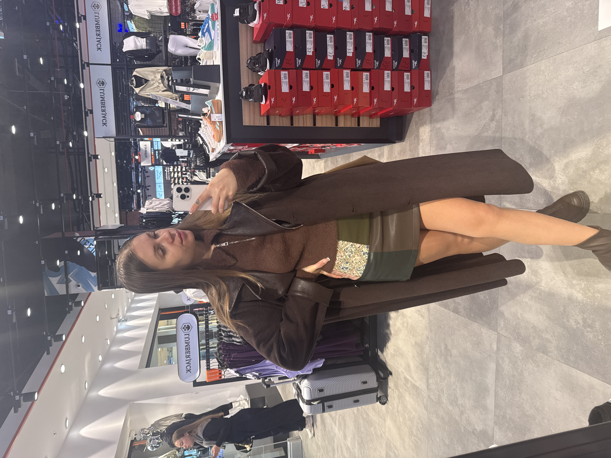 A woman is sitting on a bench in a Timberland store in Batumi, Georgia. She is wearing a brown coat, brown sweater, green leather skirt, and brown boots. She is holding a smartphone and taking a selfie. Next to her on the floor is a gray suitcase. There are clothing racks and shoe displays visible in the background.