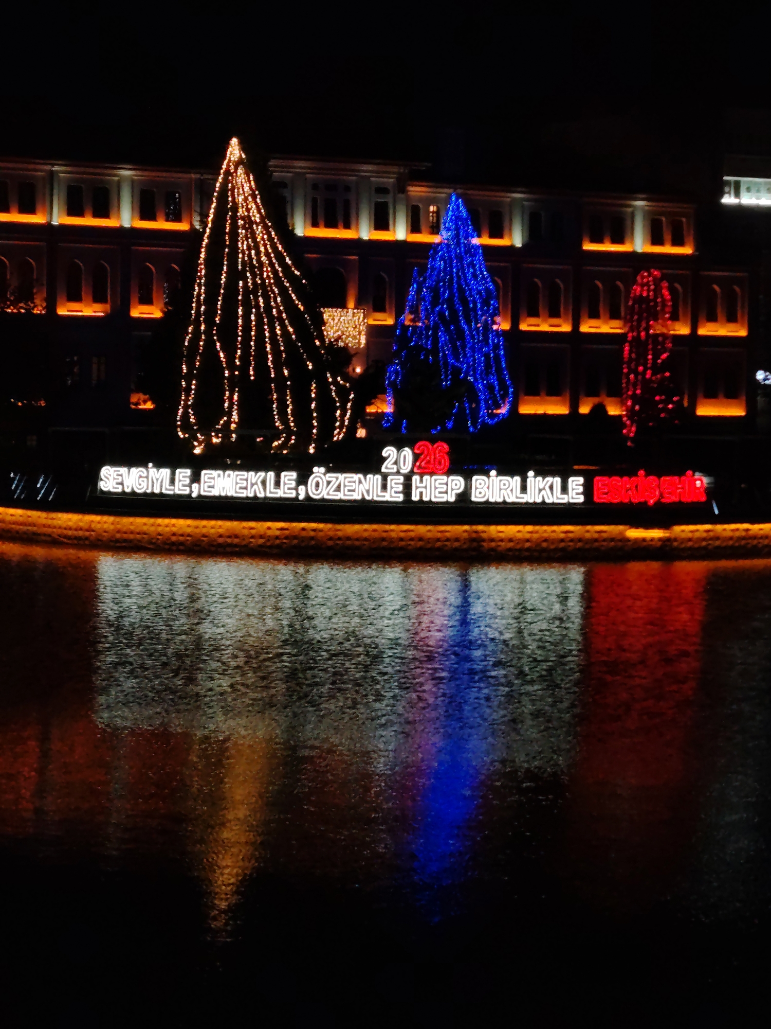 This media file captures a vibrant night scene in Eskisehir, Turkey, showcasing a festive illumination, likely along a river or canal. The foreground features the dark, rippled surface of water, which beautifully reflects the bright lights from above.
In the middle ground, three prominent, conical structures, resembling decorated trees, are covered in string lights. From left to right, these lights are golden-orange, vibrant blue, and red, creating a striking color contrast against the dark surroundings. Below these illuminated structures, a long sign displays glowing text.
In the background, a large building with multiple stories and numerous windows is brightly lit with warm orange and white lights, outlining its facade and windows and adding to the overall luminous atmosphere. The sky above is completely dark, emphasizing the brightness of the artificial lights.
The visible text on the sign reads "2026" above the main phrase "SEVGİYLE, EMEKLE, ÖZENLE HEP BİRLİKLE ESKİŞEHİR". This translates to "WITH LOVE, LABOR, CARE, ALWAYS TOGETHER ESKİŞEHİR," suggesting a message of unity and well wishes, possibly for the upcoming year 2026. The reflections of all these lights are clearly visible in the water, adding depth and a dynamic quality to the image. The scene suggests a public holiday or special event display.