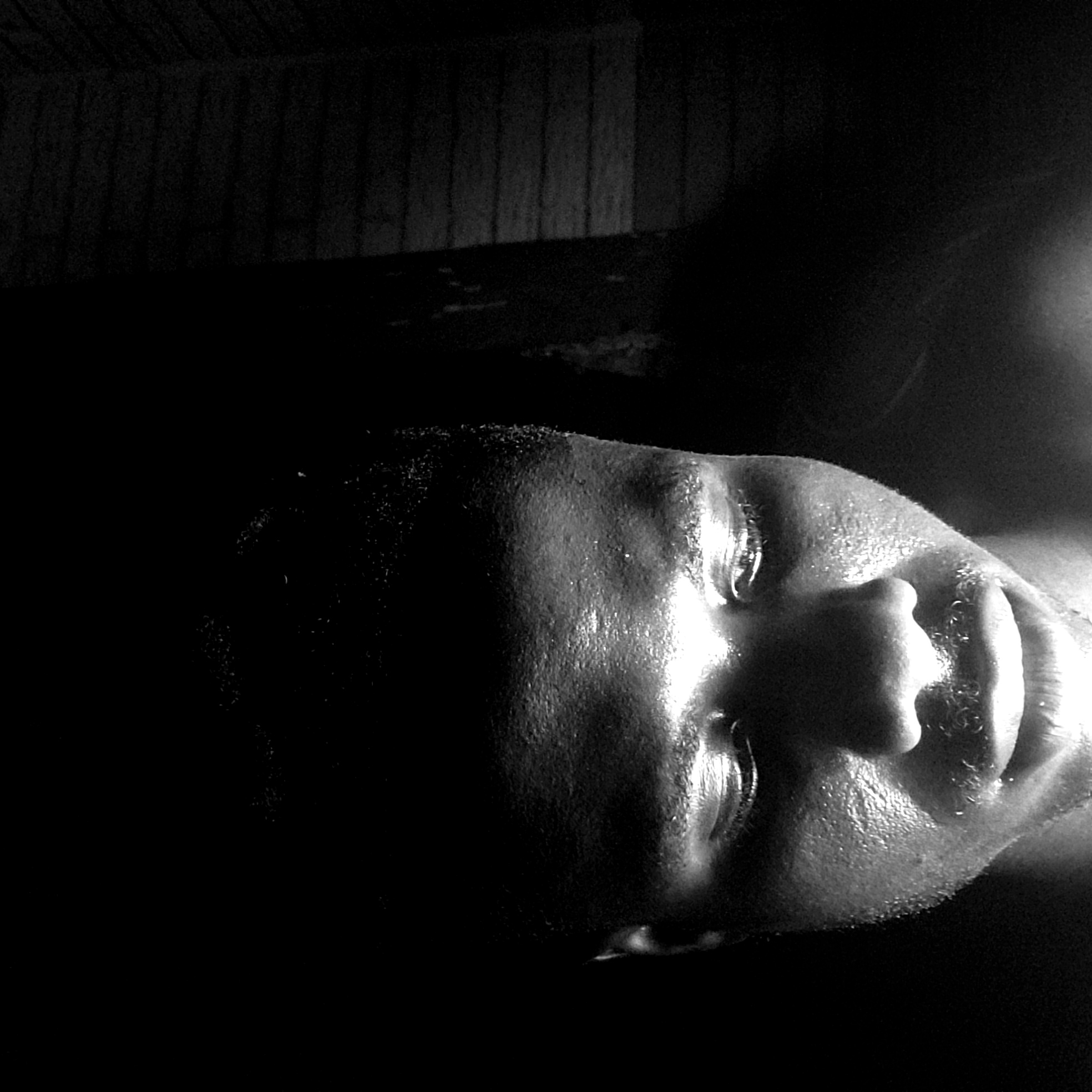 A close-up, high-contrast, black-and-white photograph depicts a person's face. The face is positioned in the foreground and appears to be slightly turned upwards. The skin texture is visible, with details highlighting the pores and facial hair. The eyes are partially open. The background is dark, with wood slats. The scene does not depict any identifiable location in Narayi, Nigeria. The focus is strictly on the person's face. No actions or interactions are observable.