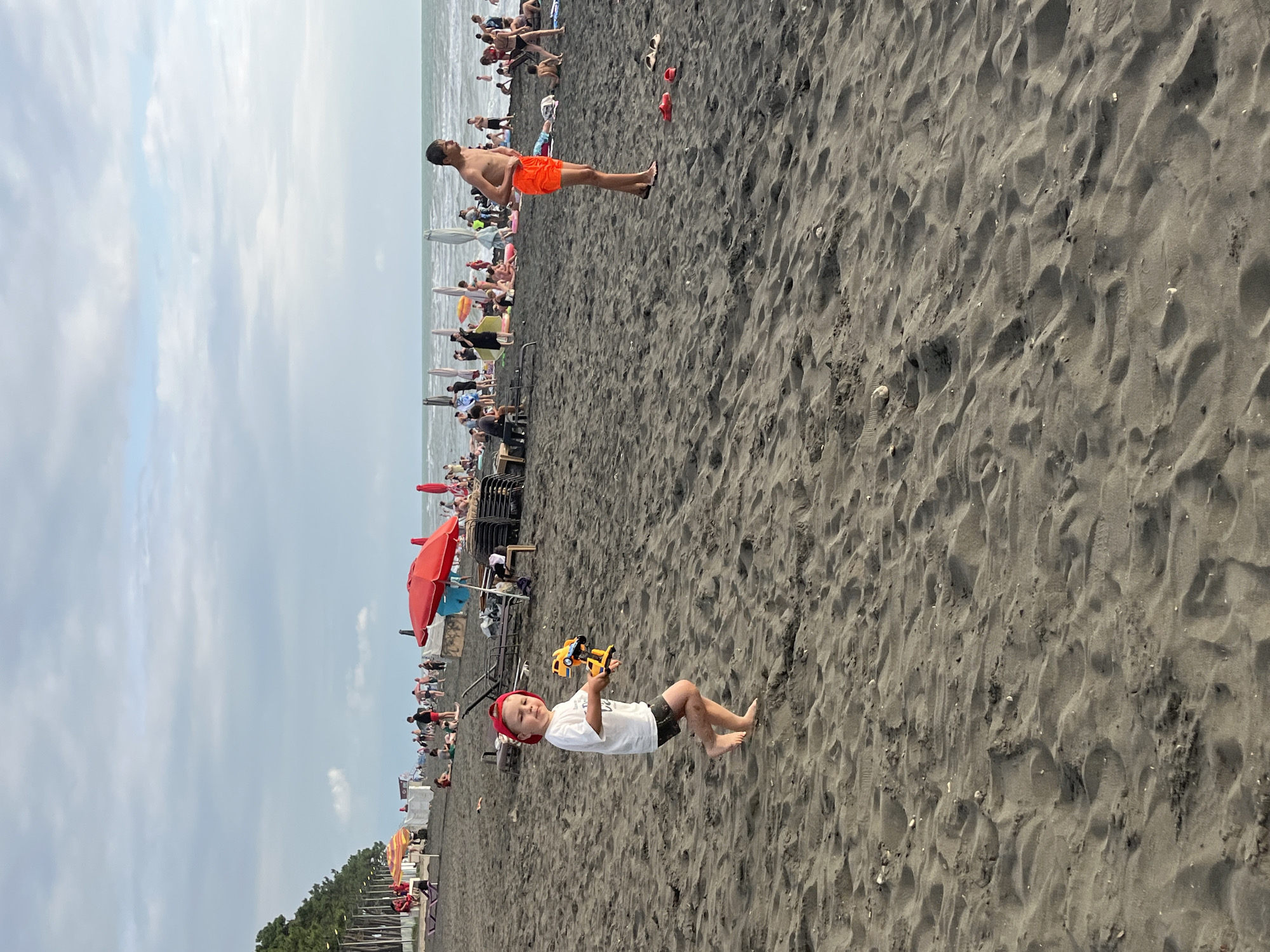 A child in a red cap and white t-shirt runs on a dark sand beach in Ureki, Georgia, holding a yellow toy. Behind the child, people relax under umbrellas, swim in the sea, and walk along the shore. In the background, a line of trees borders the beach. The sky is cloudy with patches of blue.