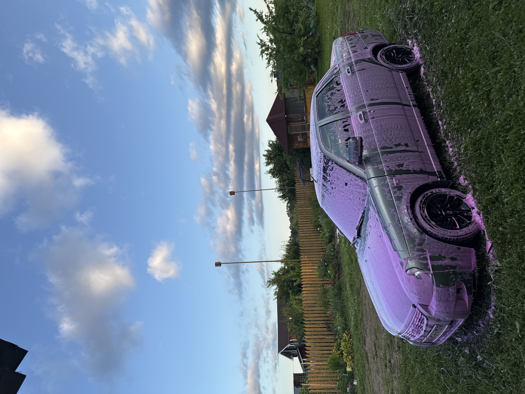 A dark-colored sedan, covered in pink foam, is parked on a grassy area in front of a wooden fence and a small, rustic gazebo. The car, likely undergoing a car wash, has black rims on its wheels. Behind the fence, a wooden house with a dark roof and lush green trees are visible. A tall, thin pole with a small wooden structure on top stands near the fence. The sky above is a bright blue with scattered white and gray clouds, suggesting it is either early morning or late afternoon. The overall setting appears to be a rural or suburban backyard in Vereshchagino, Russia.